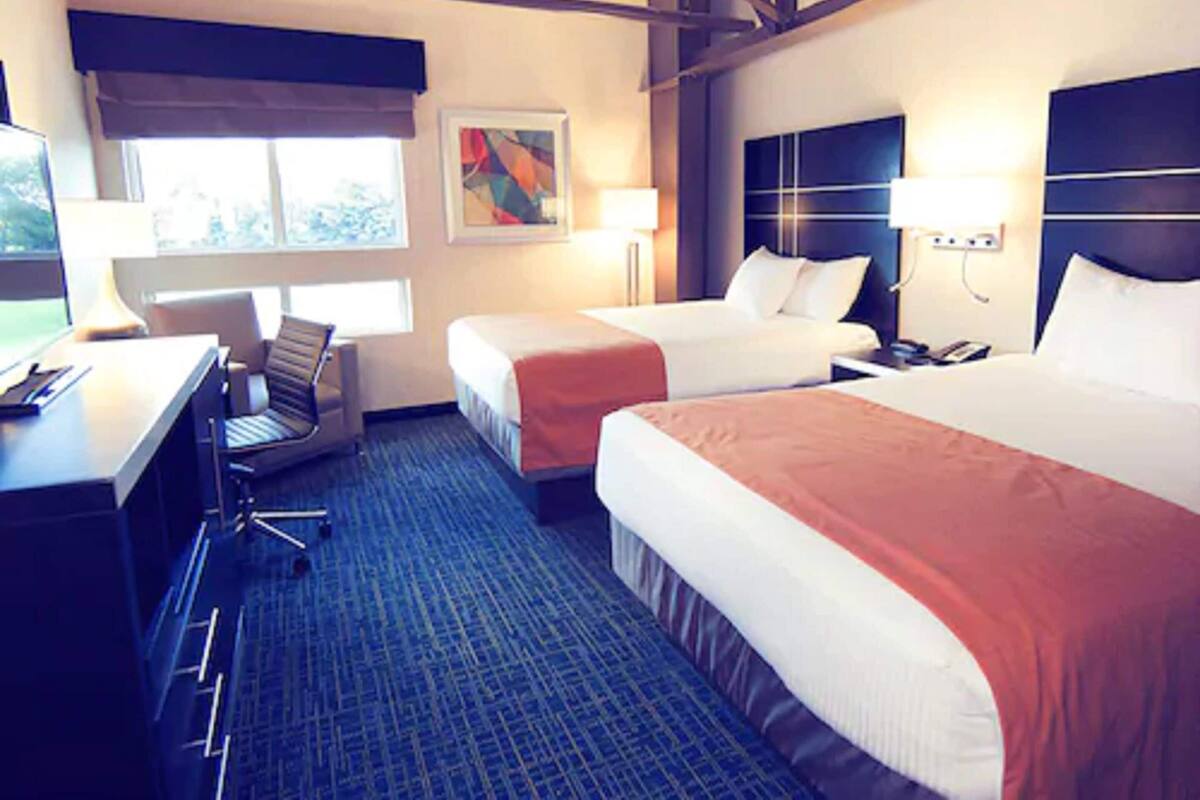 A hotel room with two beds, a desk, a chair, a lamp, and a painting on the wall.