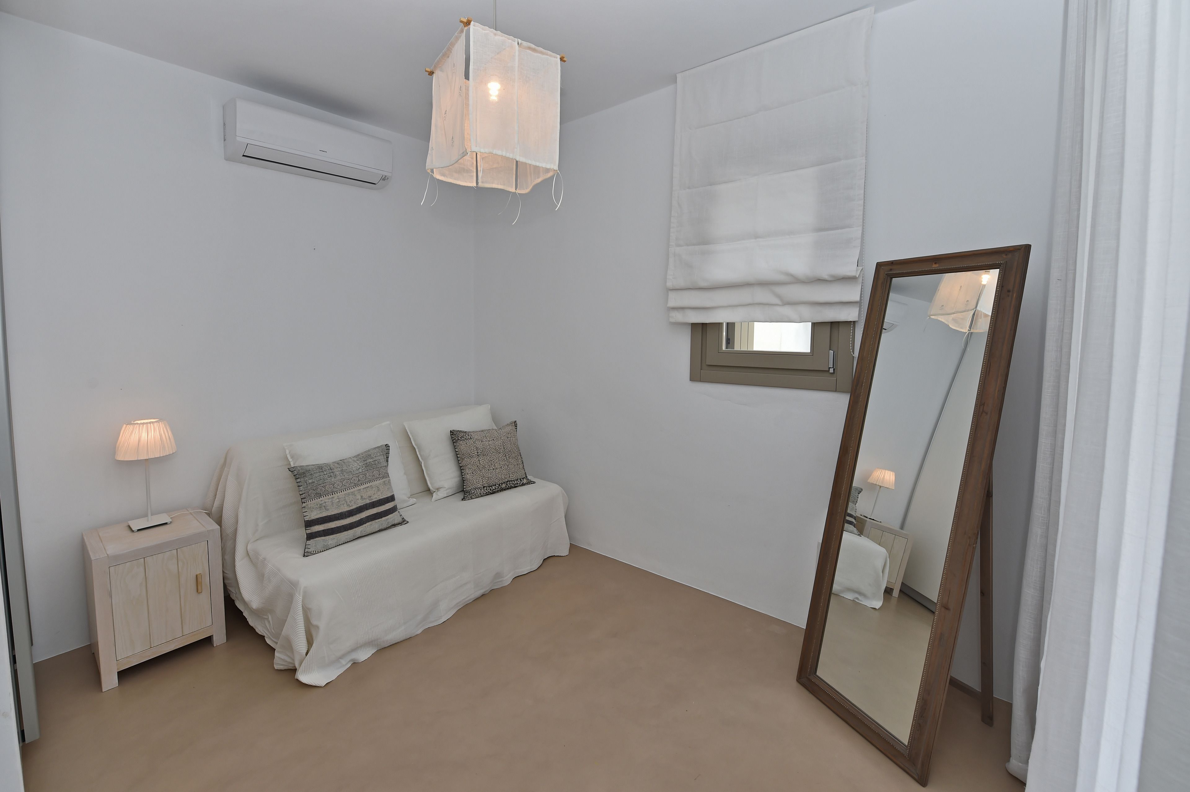 A bedroom with a bed, bedside table, lamp, air conditioning unit, window with blinds, and a mirror.