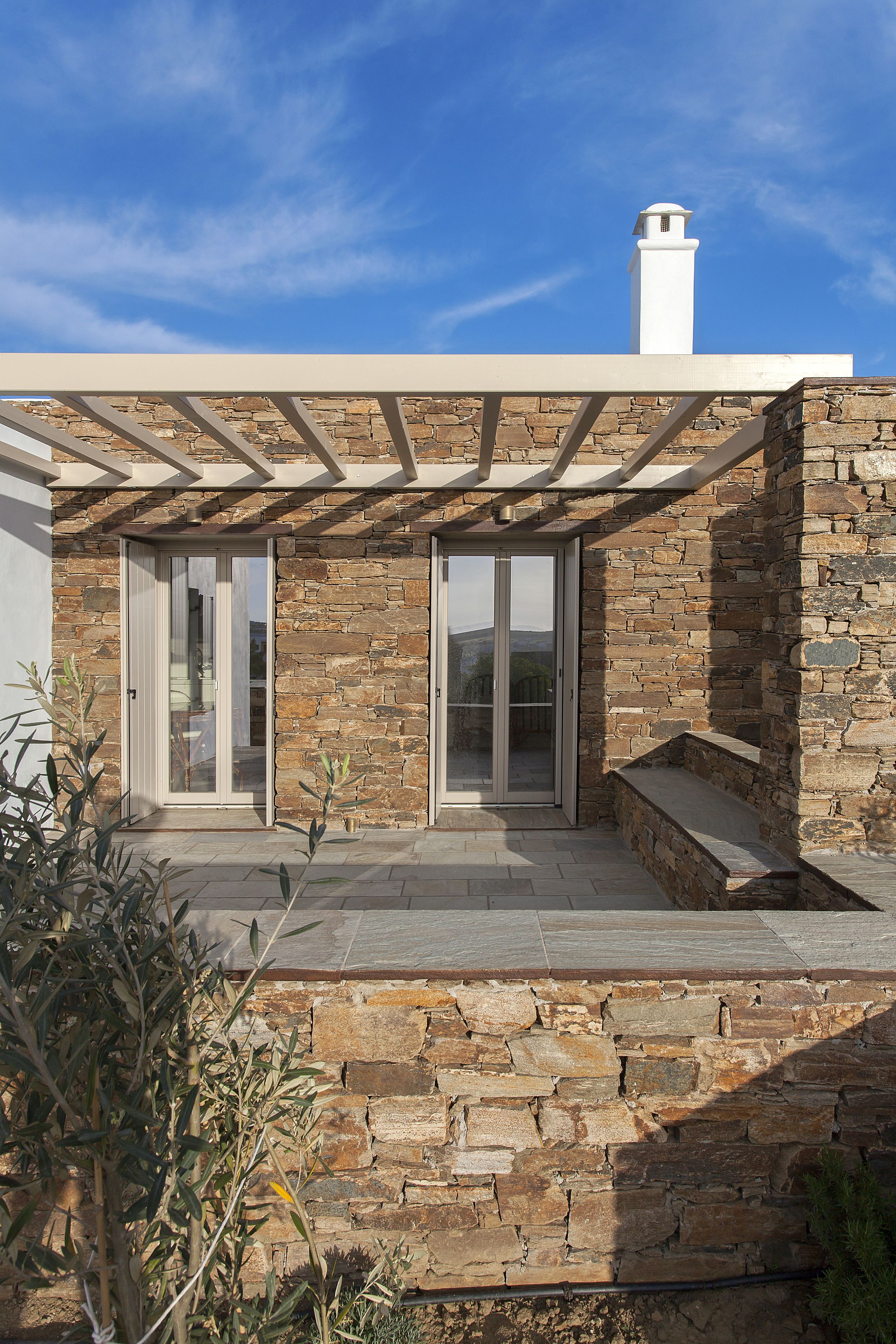 Une maison moderne aux murs en pierre, dotée d’une cheminée blanche et d’un patio couvert avec des portes-fenêtres coulissantes.
