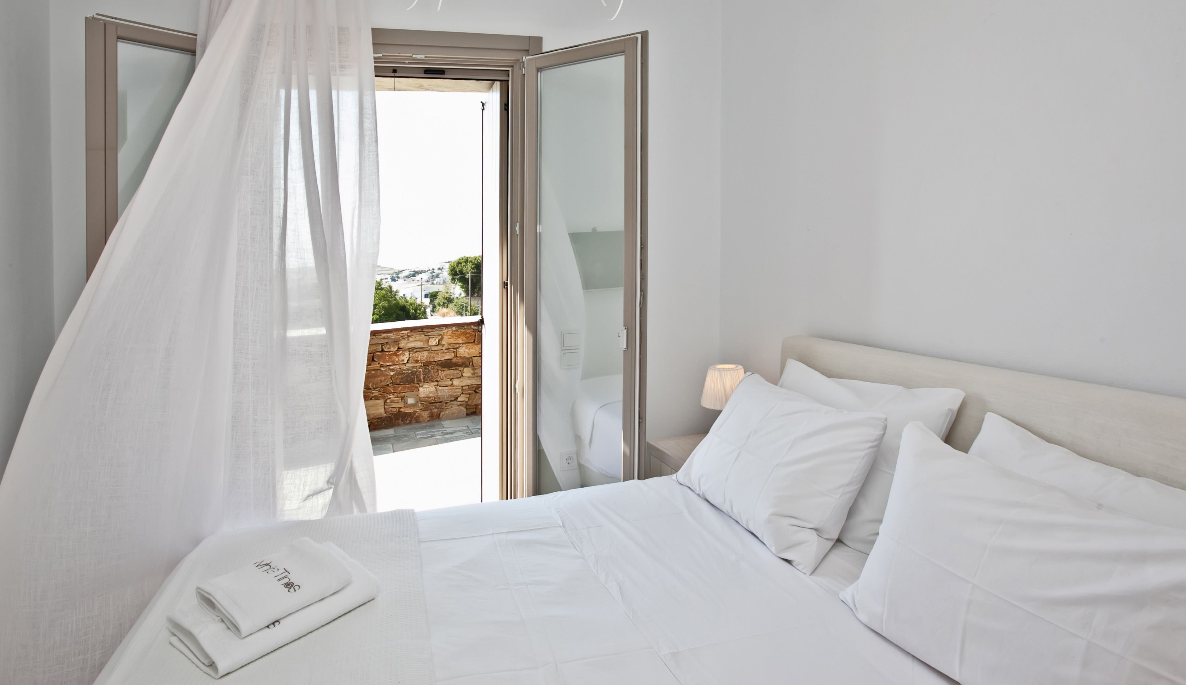 A bedroom with a bed, white bedding, a bedside table with a lamp, and a view of a stone wall and greenery outside.