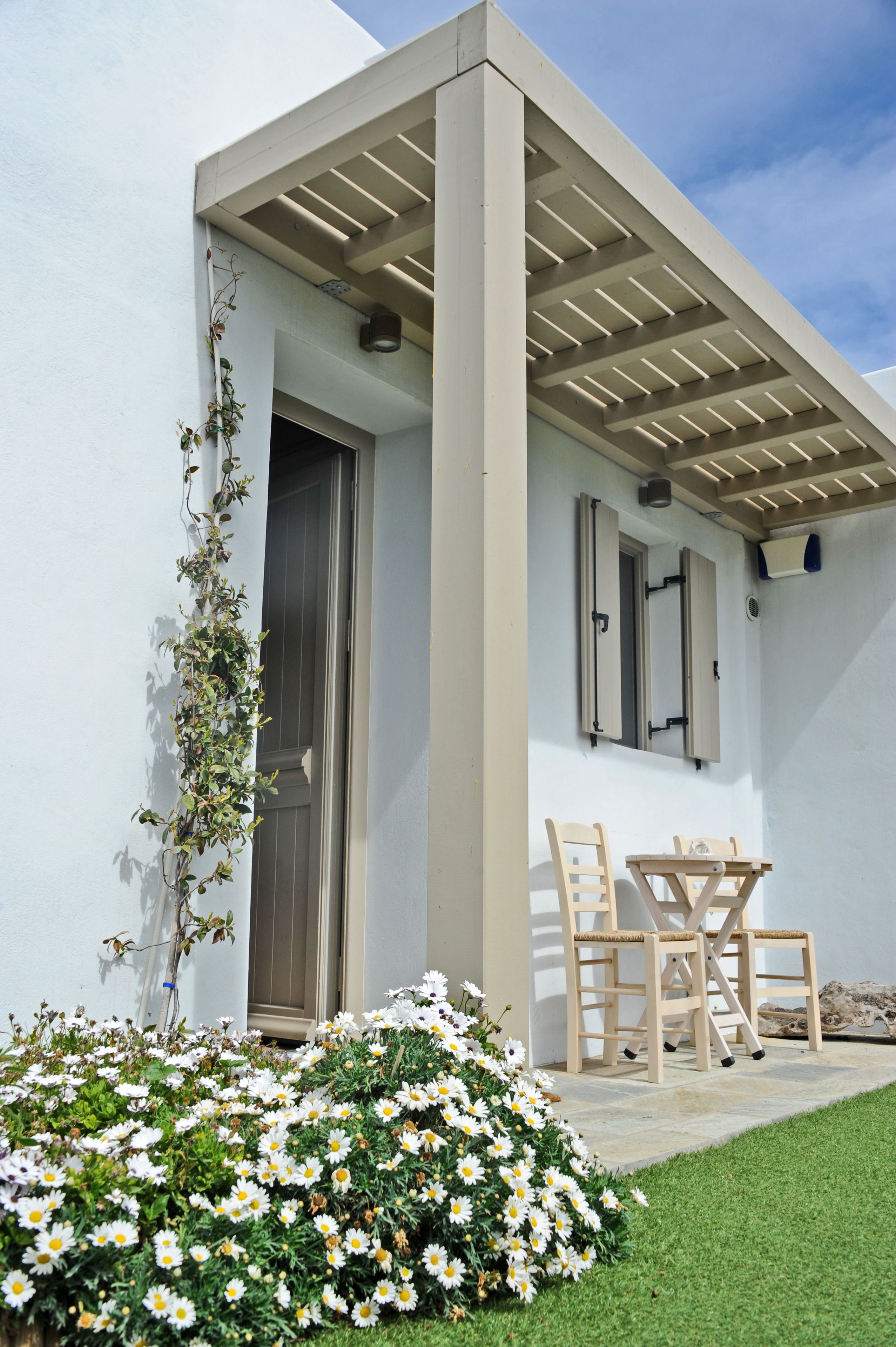 Ein modernes Gebäude mit weißer Fassade, einer Holzpergola, einer Tür und einem kleinen Tisch mit Stühlen.
