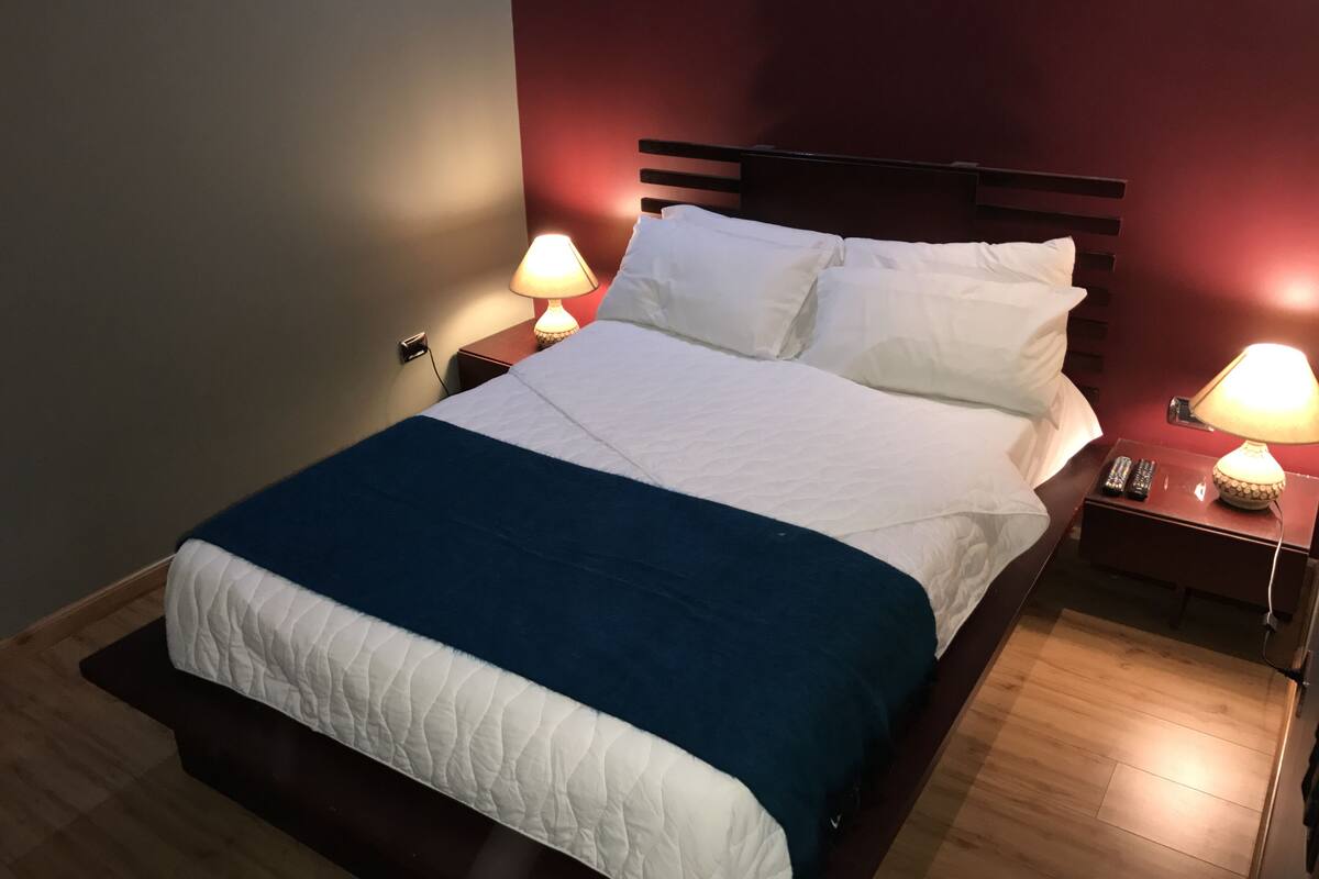 A neatly made bed with white and blue bedding, two bedside lamps, and a wooden nightstand with a remote control.