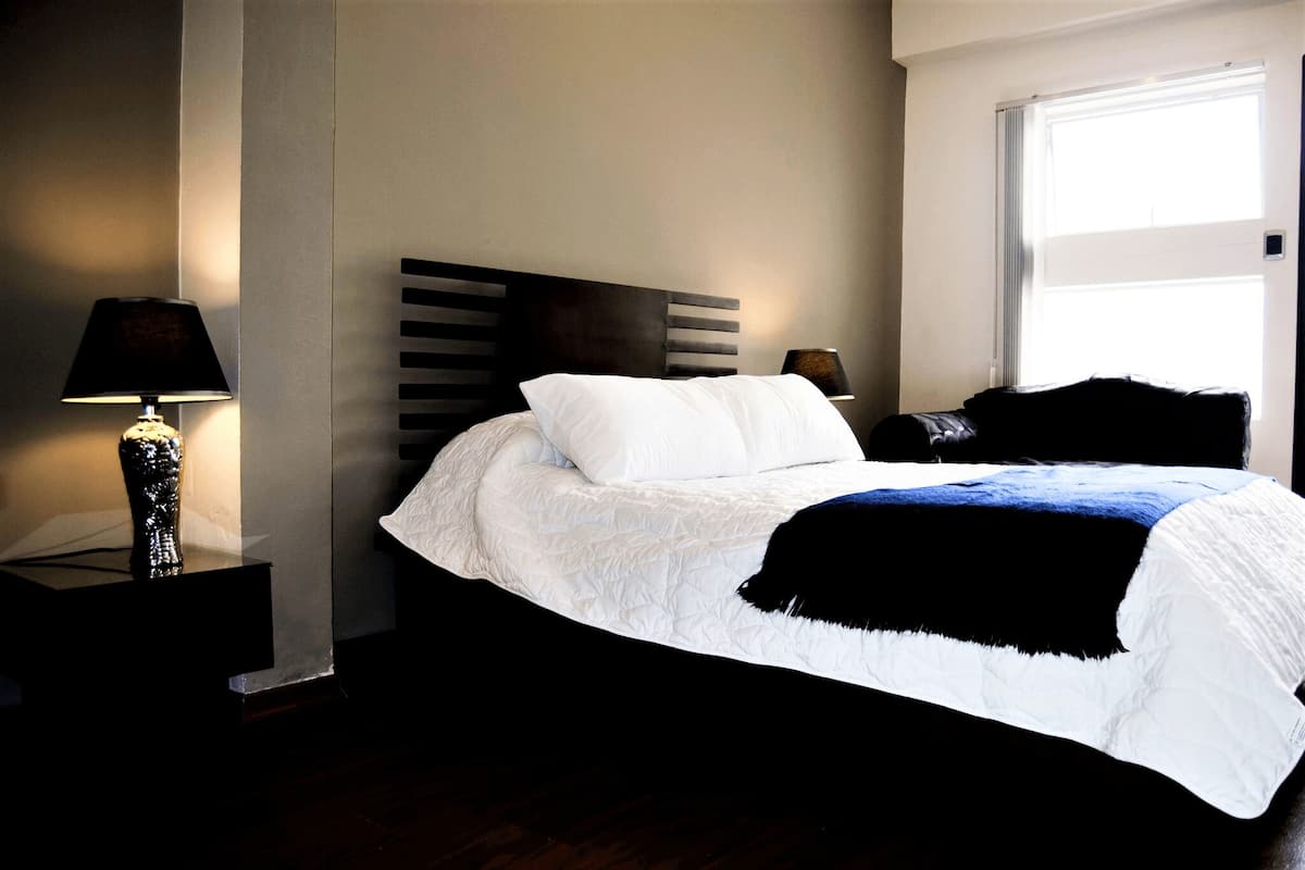 A neatly made bed with white bedding and a black throw blanket, a bedside lamp, and a window with blinds.