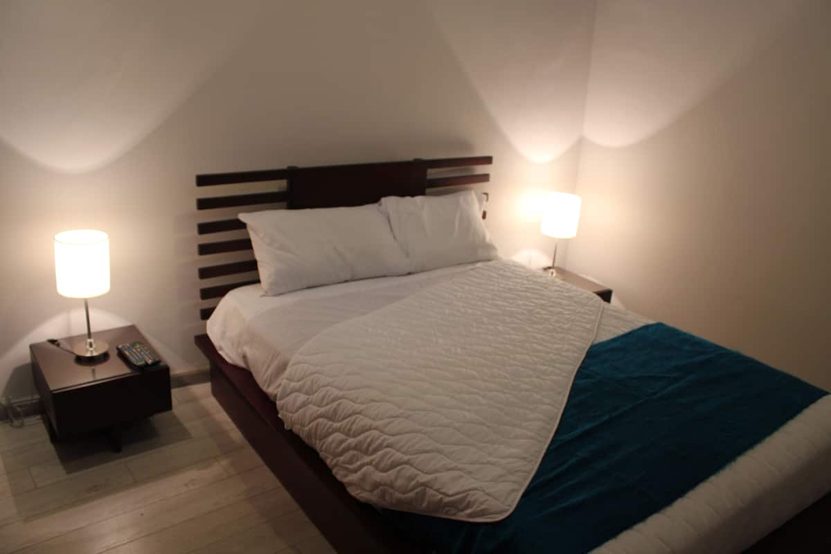 A neatly made bed with a white quilt and a teal throw blanket, flanked by two bedside tables with lamps, in a room with a wooden headboard and white walls.