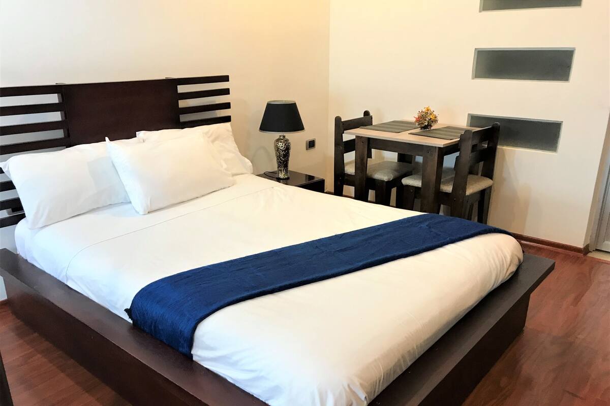 A neatly made bed with white linens and a blue accent, a bedside table with a lamp, and a small table with a vase of flowers.