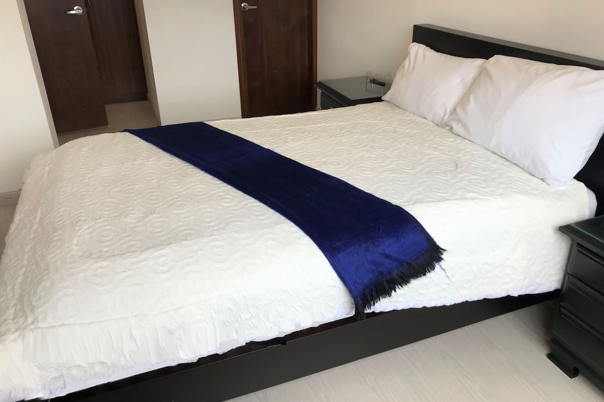 A neatly made bed with a white and blue bedspread, a dark headboard, and a nightstand with a lamp.