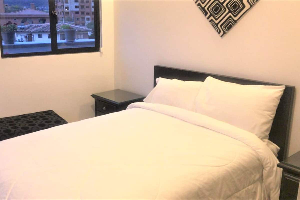 A neatly made bed with white linens, a small bedside table, and a window with a view of buildings.
