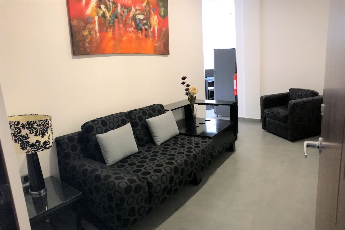 A living room with a patterned sofa, a side table with a lamp, a small table with a vase, and a black armchair.