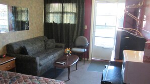 A room with a sofa, a small table with a vase of flowers, a chair, a television, and a sliding door leading to an outdoor area. - The New Elgin Motel (Saint Thomas)