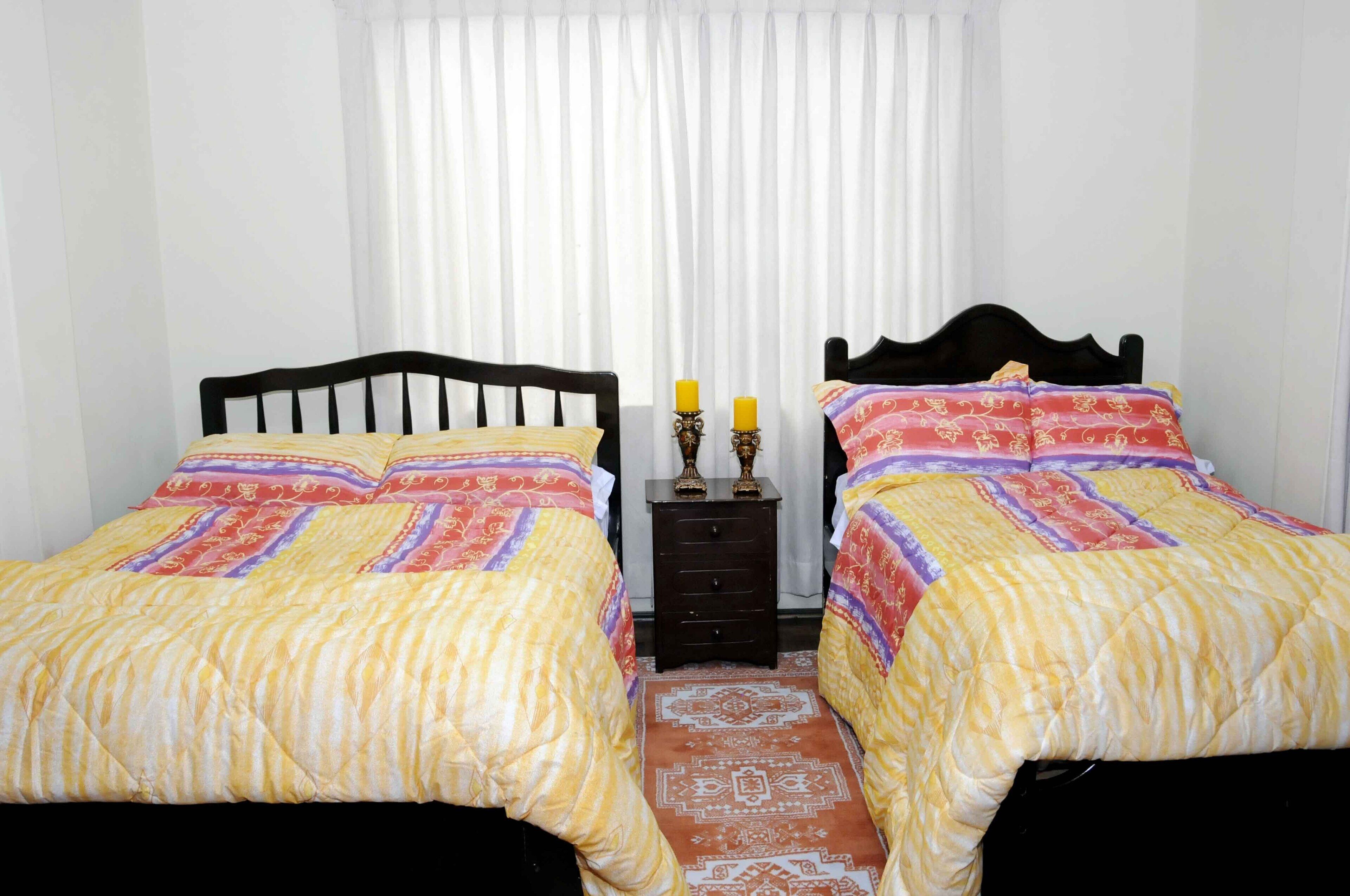 Una habitación con dos camas, cada una con cabecera y colcha estampada. Entre las camas, una mesita de noche de madera con dos velas.