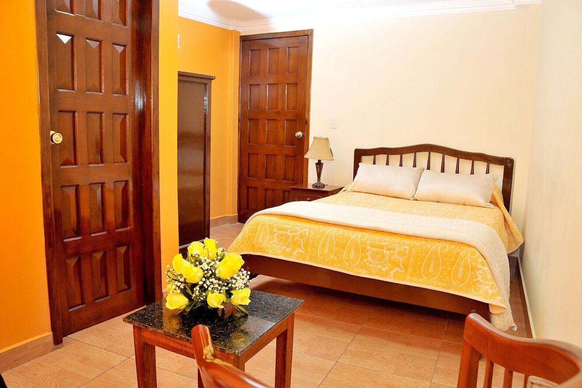 A bedroom with a bed, a nightstand, a lamp, a table with a vase of flowers, and two wooden doors.