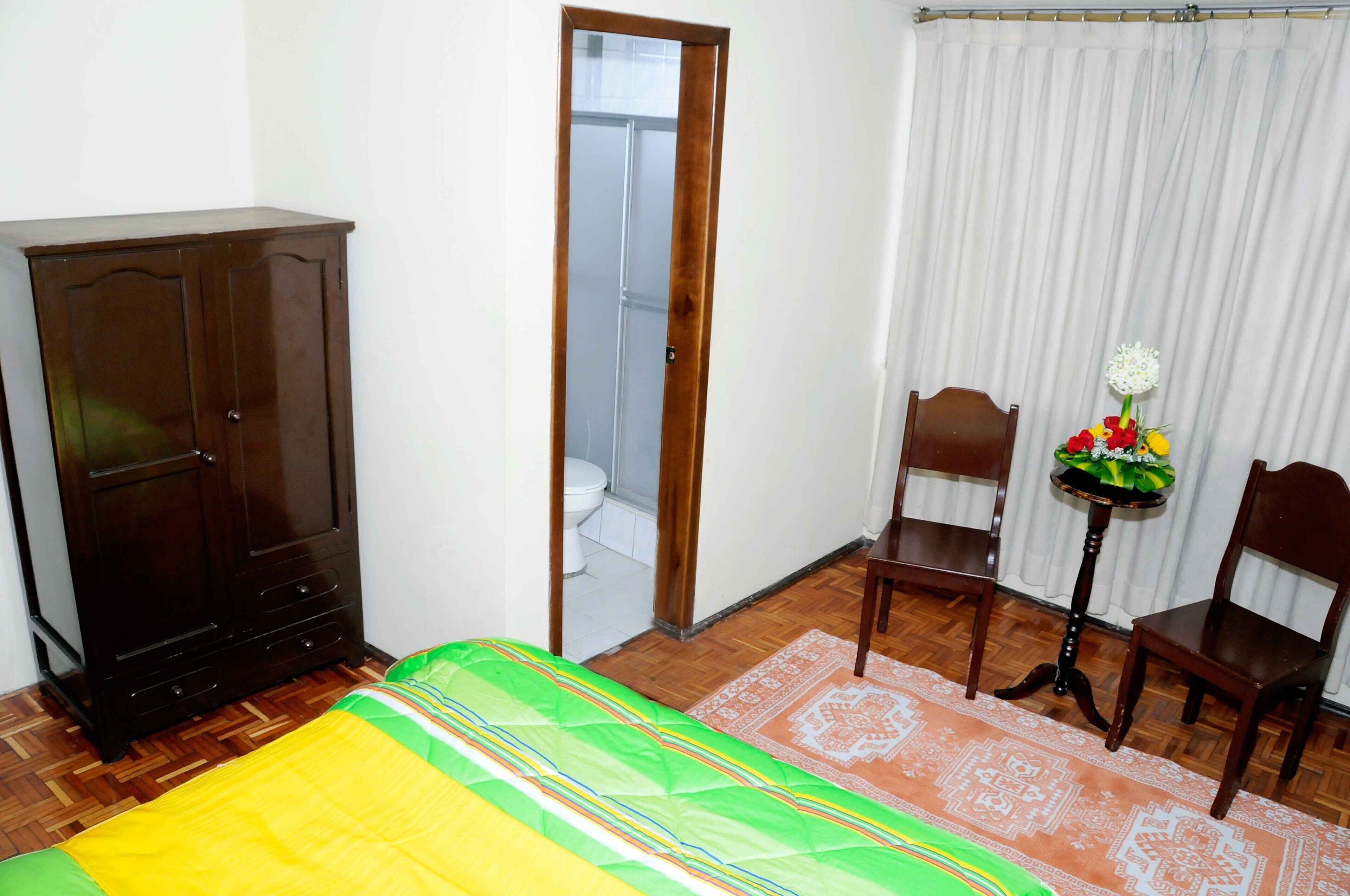 Una habitación con cama, armario, una pequeña mesa con flores, dos sillas y una cortina.
