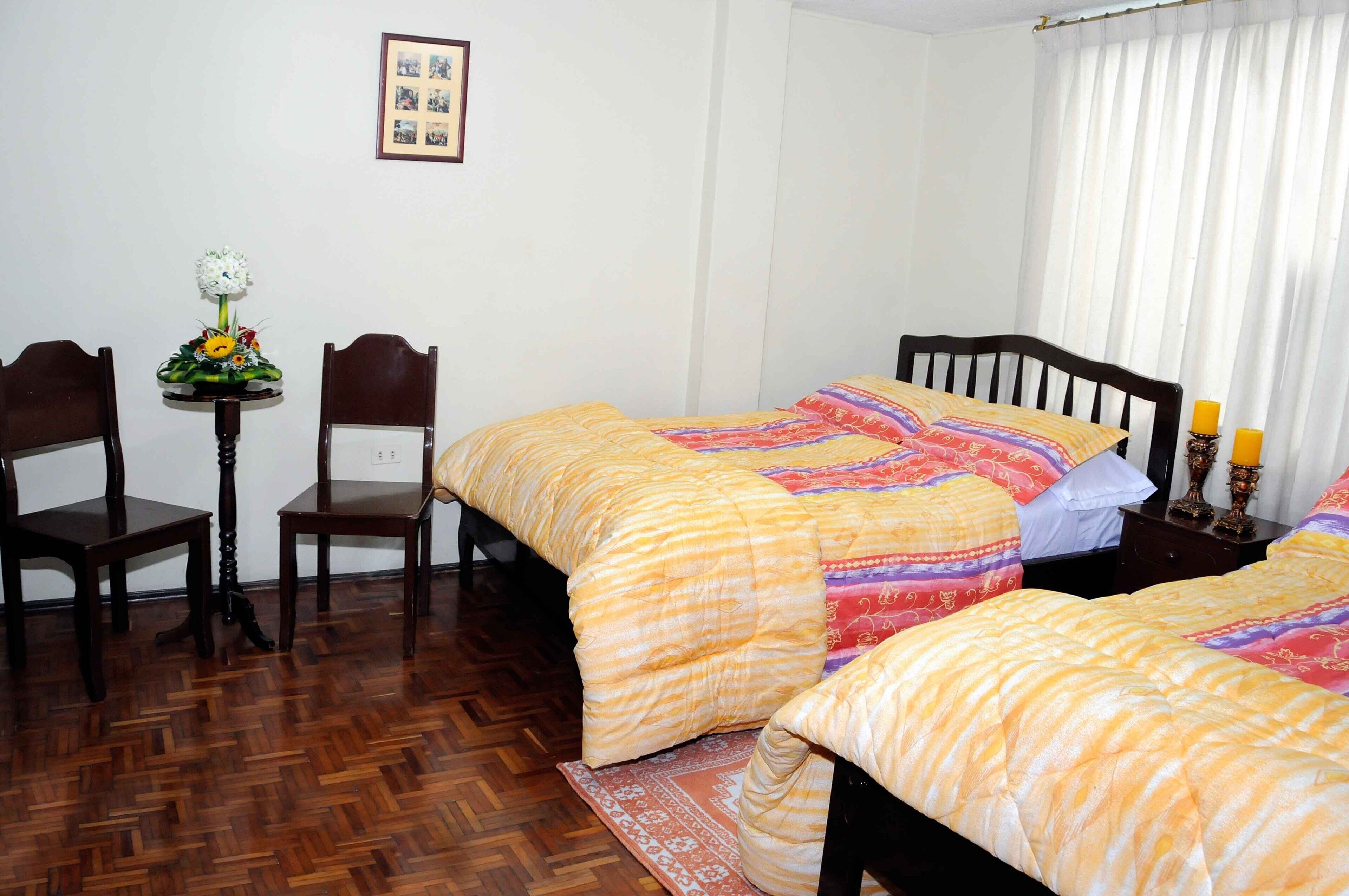 A bedroom with two beds, a wooden floor, a small table with a flower arrangement, and a framed picture on the wall.