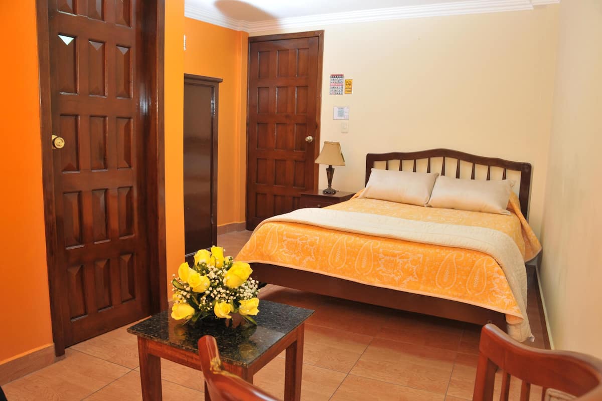 A hotel room with a bed, a wooden door, a small table with a vase of flowers, and a chair.