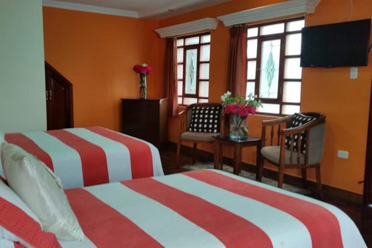 A hotel room with two beds, a desk, a chair, a TV, and a window with curtains.