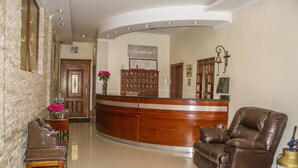 A reception area with a wooden reception desk, a brown leather sofa, a vase with flowers, and a bulletin board. - Hotel Margarita 2 (Quito)