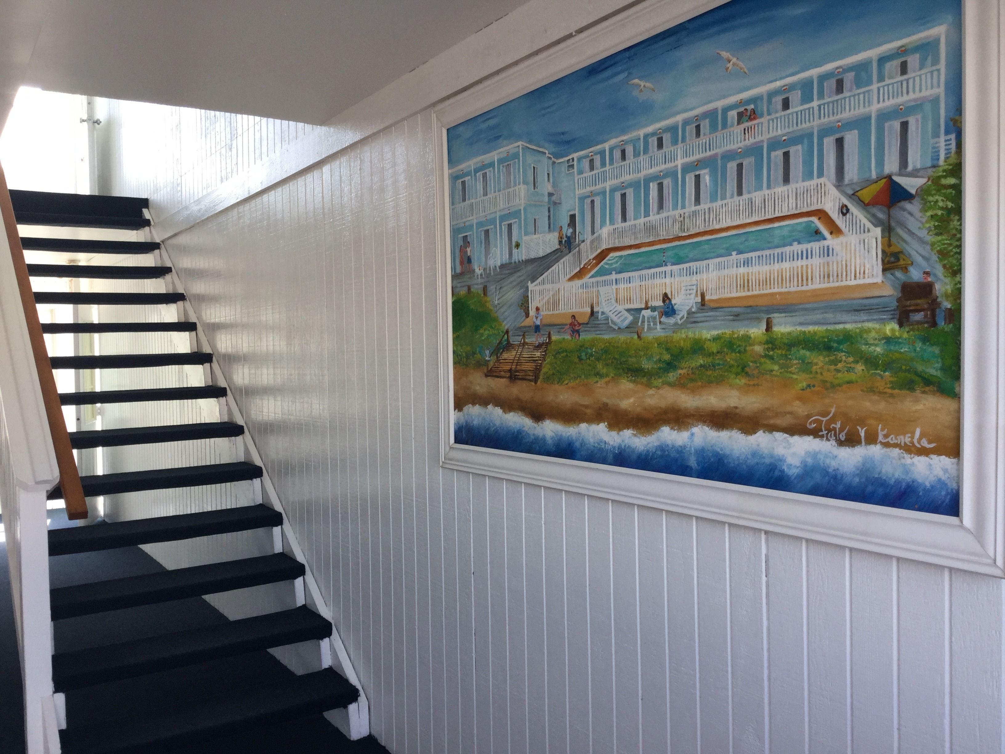 Eine Treppe mit schwarzen Stufen und weißen Wänden, die zu einem Raum führt, der ein Wandgemälde eines blauen Gebäudes und einer Strandansicht zeigt.