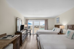 A hotel room with two beds, a desk, a television, and a balcony with a view of the sea.