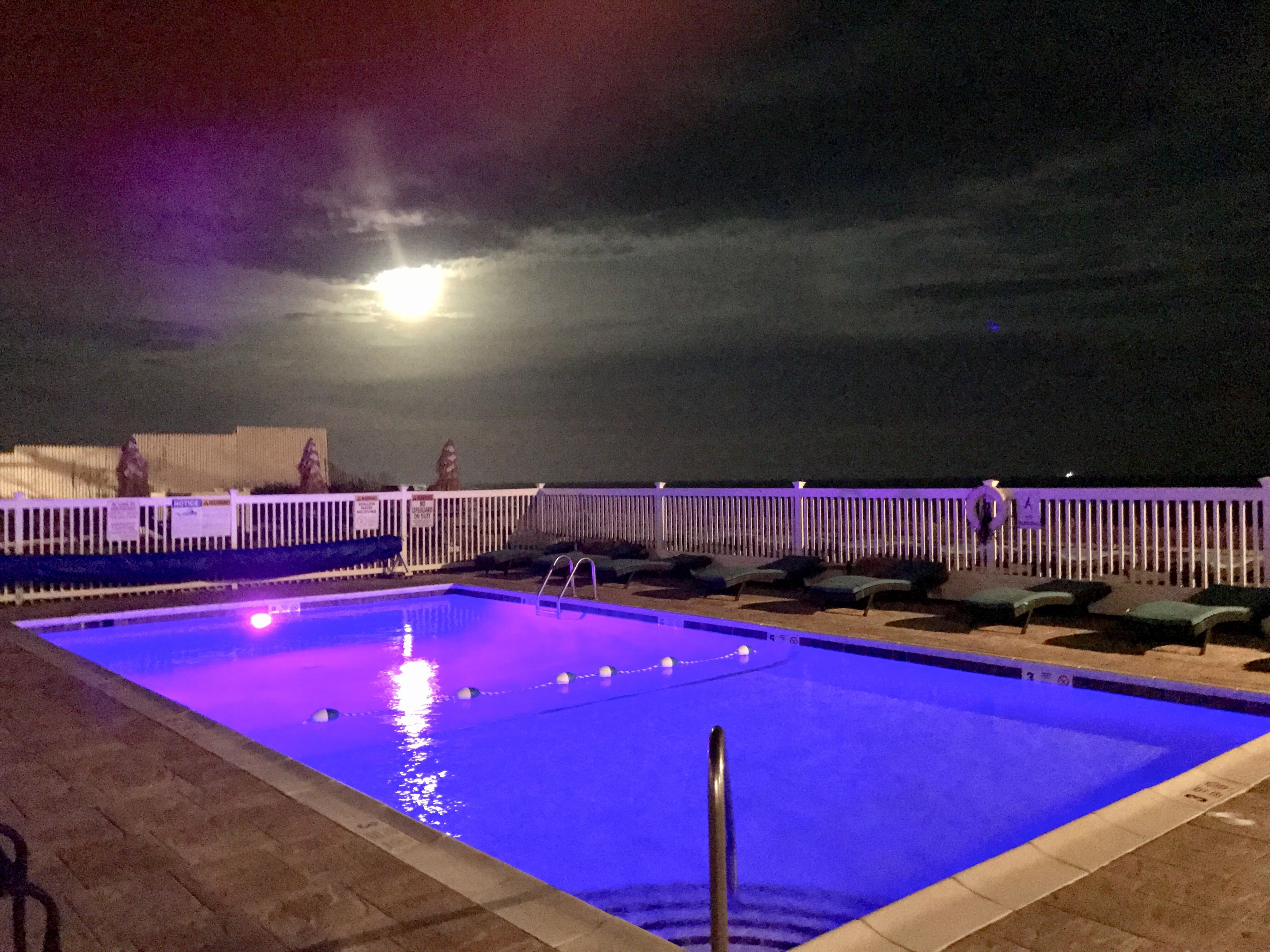 Ein Swimmingpool mit weißem Zaun und Liegestühlen, unter einem Nachthimmel mit sichtbarem Mond.