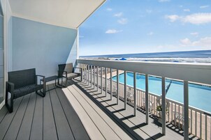 A balcony with a view of the ocean, a pool, and a seating area with chairs.