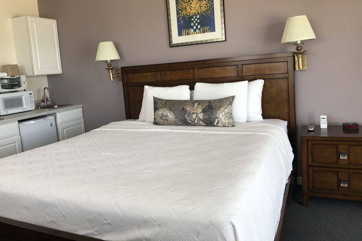 A hotel room with a bed, a nightstand, a lamp, a microwave, a small refrigerator, and a painting on the wall.