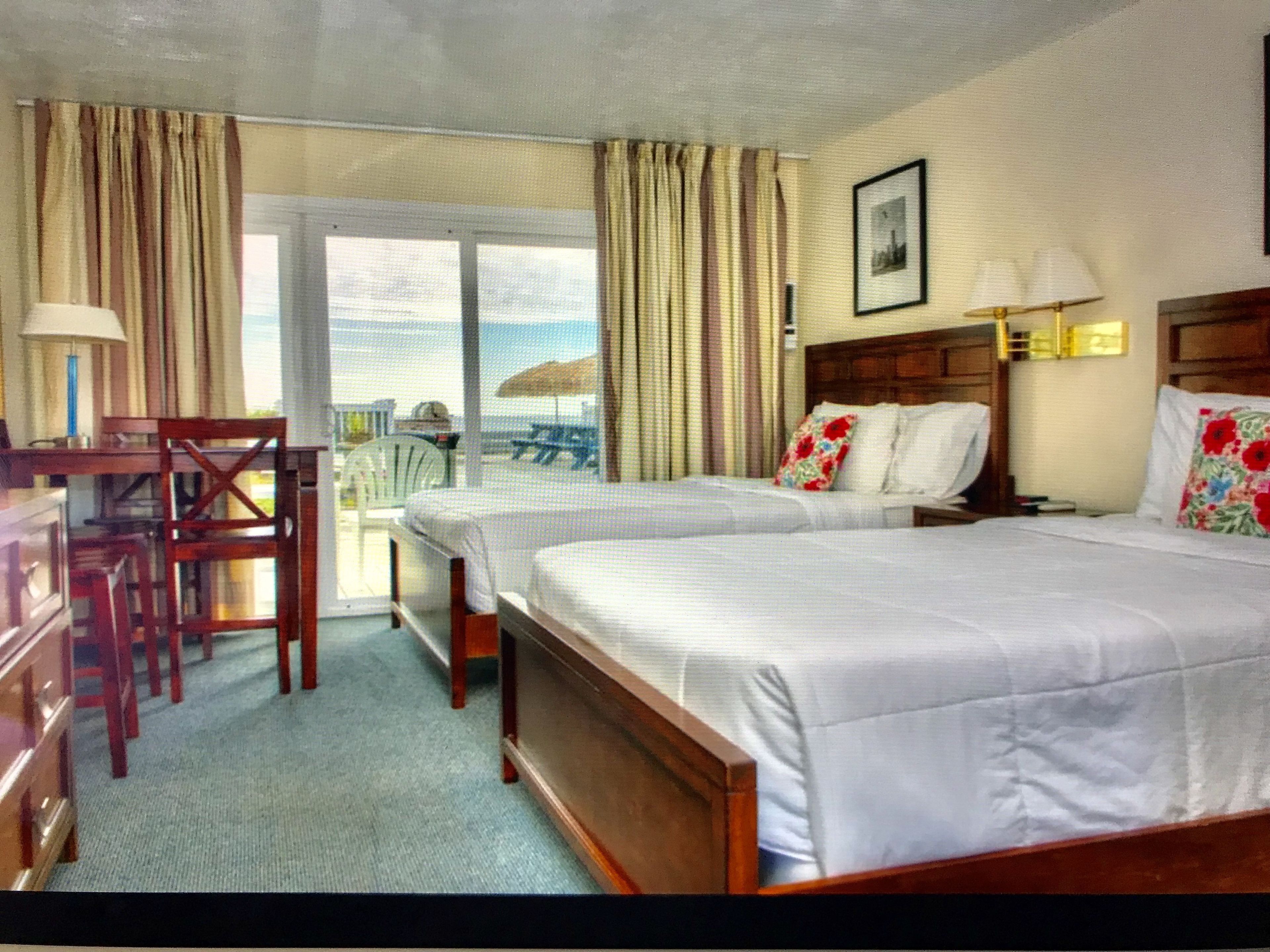 A hotel room with two beds, a desk, chairs, and a balcony with a view.