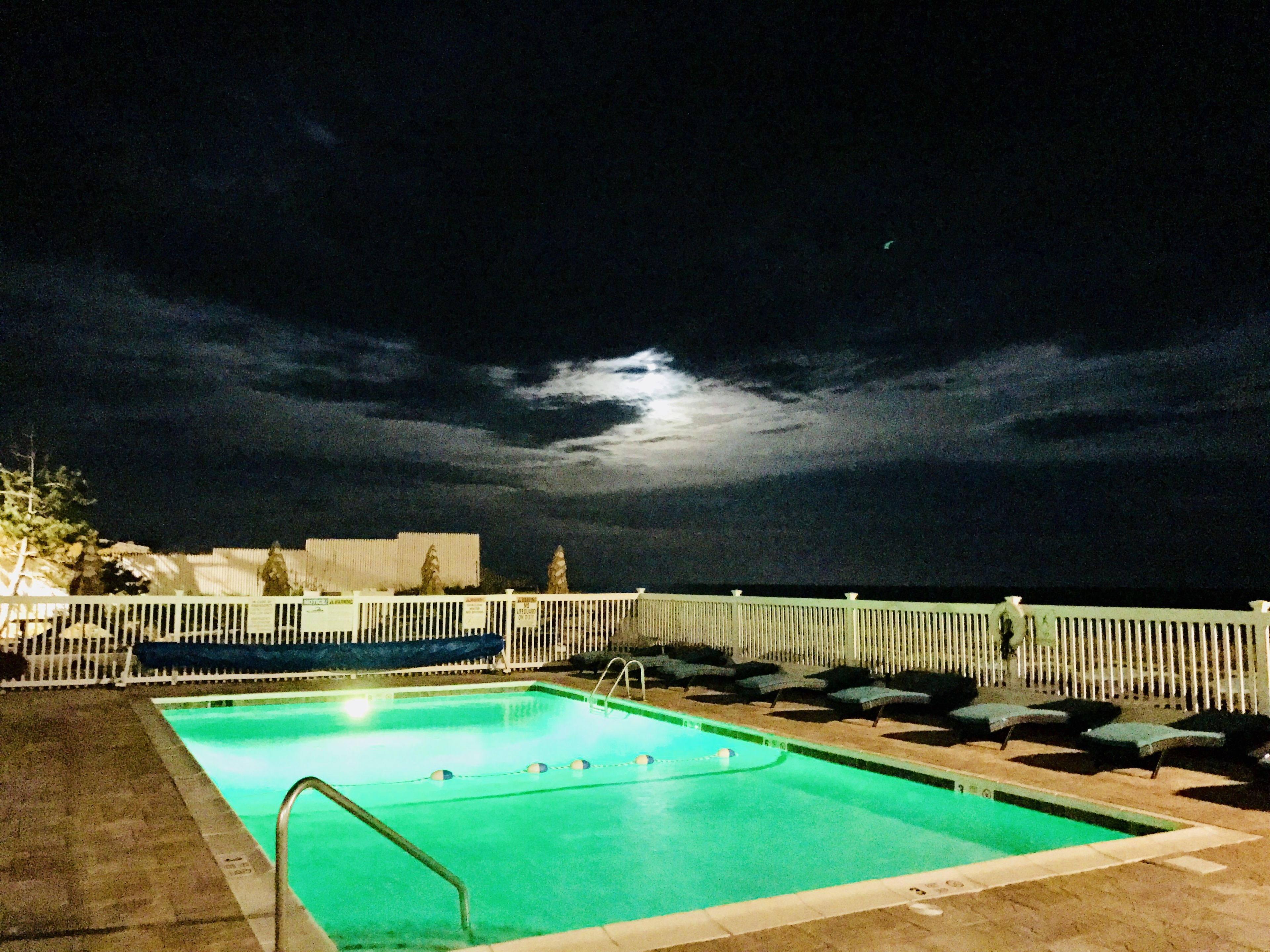 A rectangular pool with a metal ladder, surrounded by a white fence and lounge chairs, under a dark sky with a visible moon.