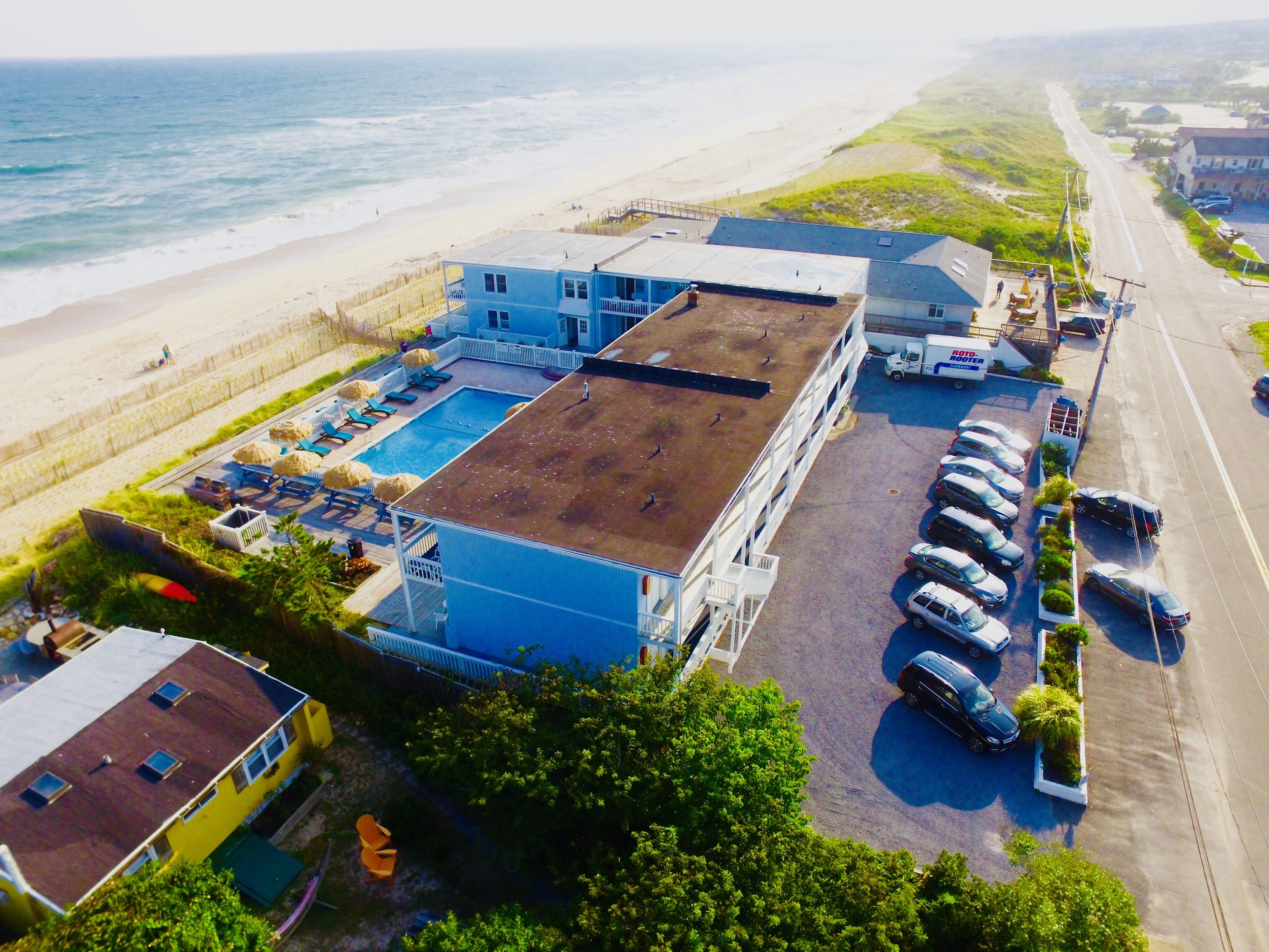 Luftaufnahme eines Gebäudes mit Schwimmbad, angrenzend an einen Strand und einen Parkplatz.