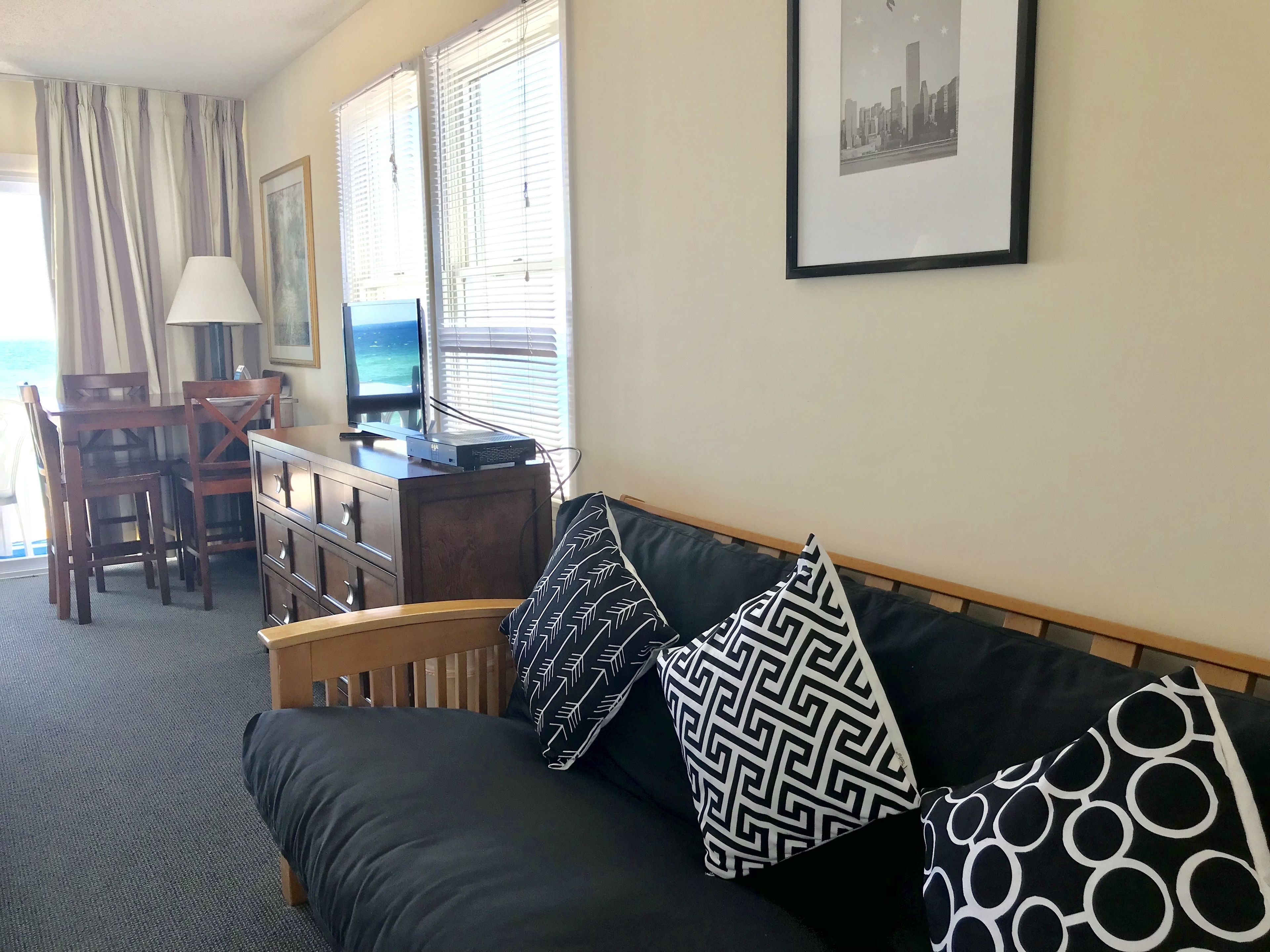 A hotel room with a dark-colored sofa, a wooden desk with a computer, a TV, and a chair. There are patterned pillows on the sofa, a lamp, and a framed picture on the wall.