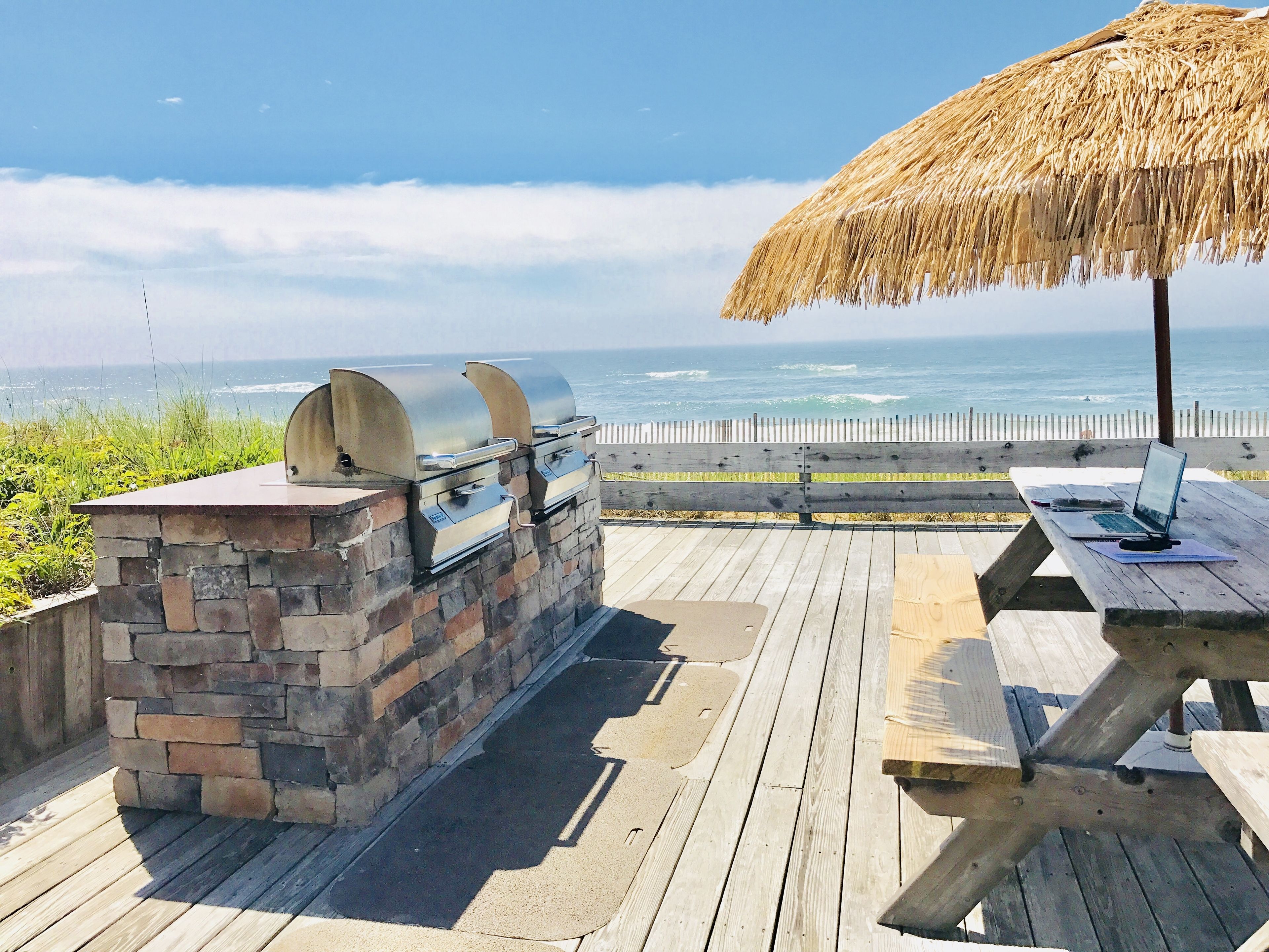 An outdoor patio with a stone barbecue grill, a wooden table with a laptop, and a thatched umbrella.