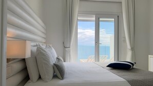 A bedroom with a bed, pillows, a bedside table, and a view of the sea through a large window. - Hôtel La Calanque (Cavalaire-sur-Mer)