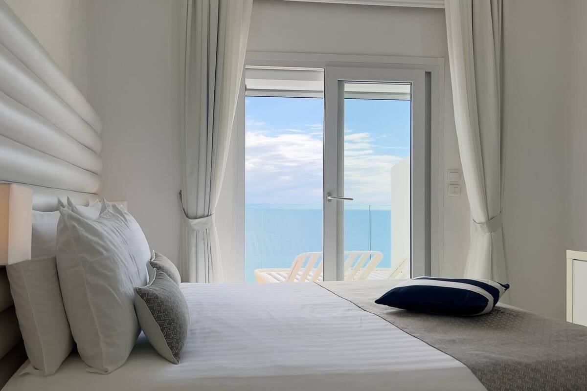 Un dormitorio con cama, almohadas, una mesita de noche y vistas al mar a través de un amplio ventanal.