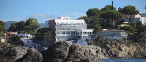 A coastal building with multiple balconies, surrounded by rocky terrain and trees.