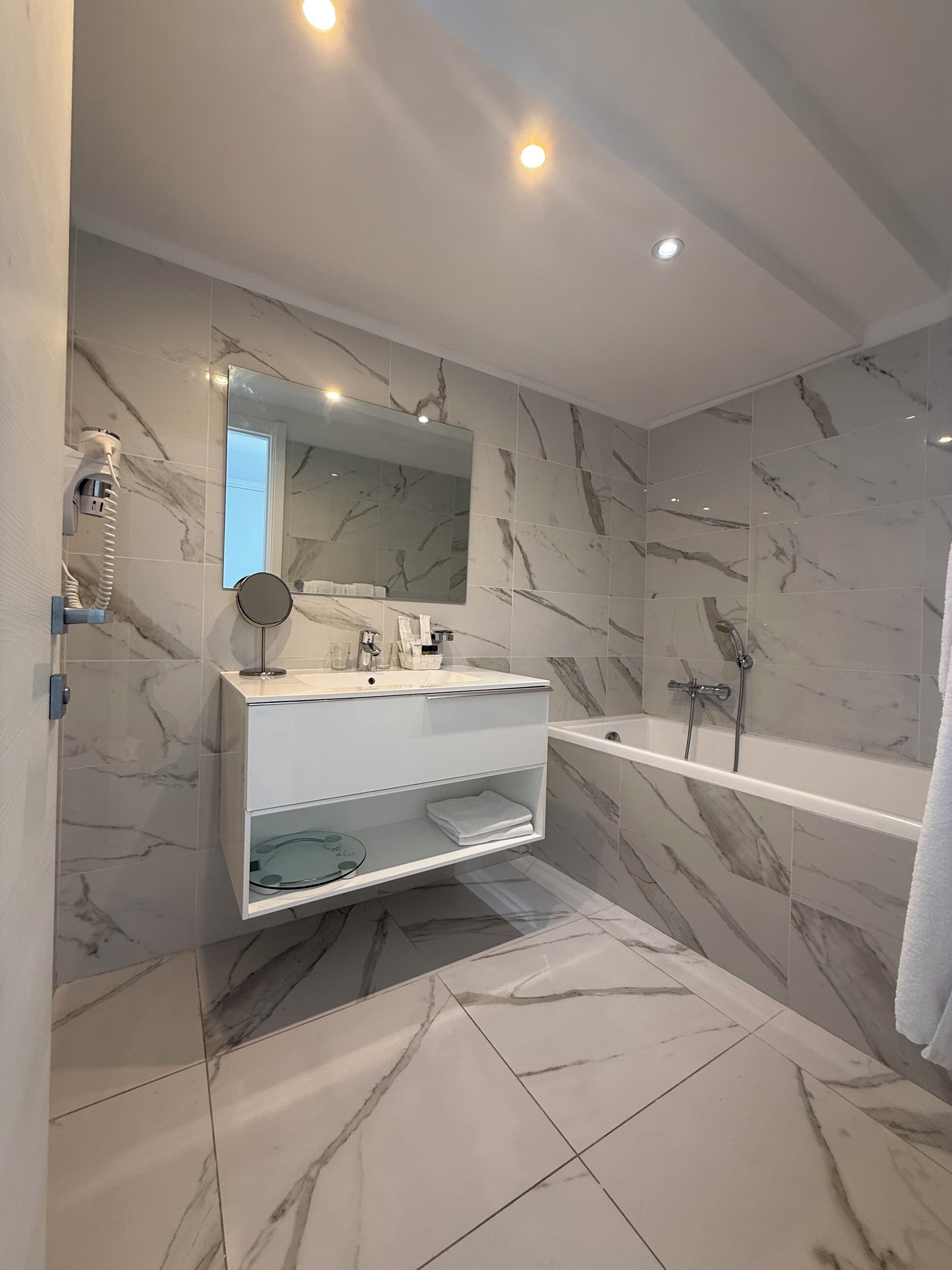 A modern bathroom with a marble-tiled floor, a white vanity with a mirror, a glass shelf, and a bathtub.