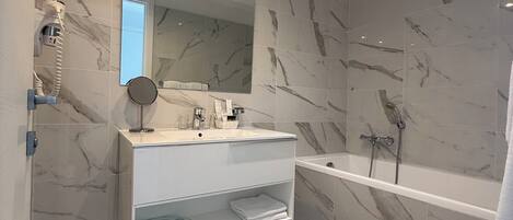 A modern bathroom with a marble-tiled floor, a white vanity with a mirror, a glass shelf, and a bathtub.