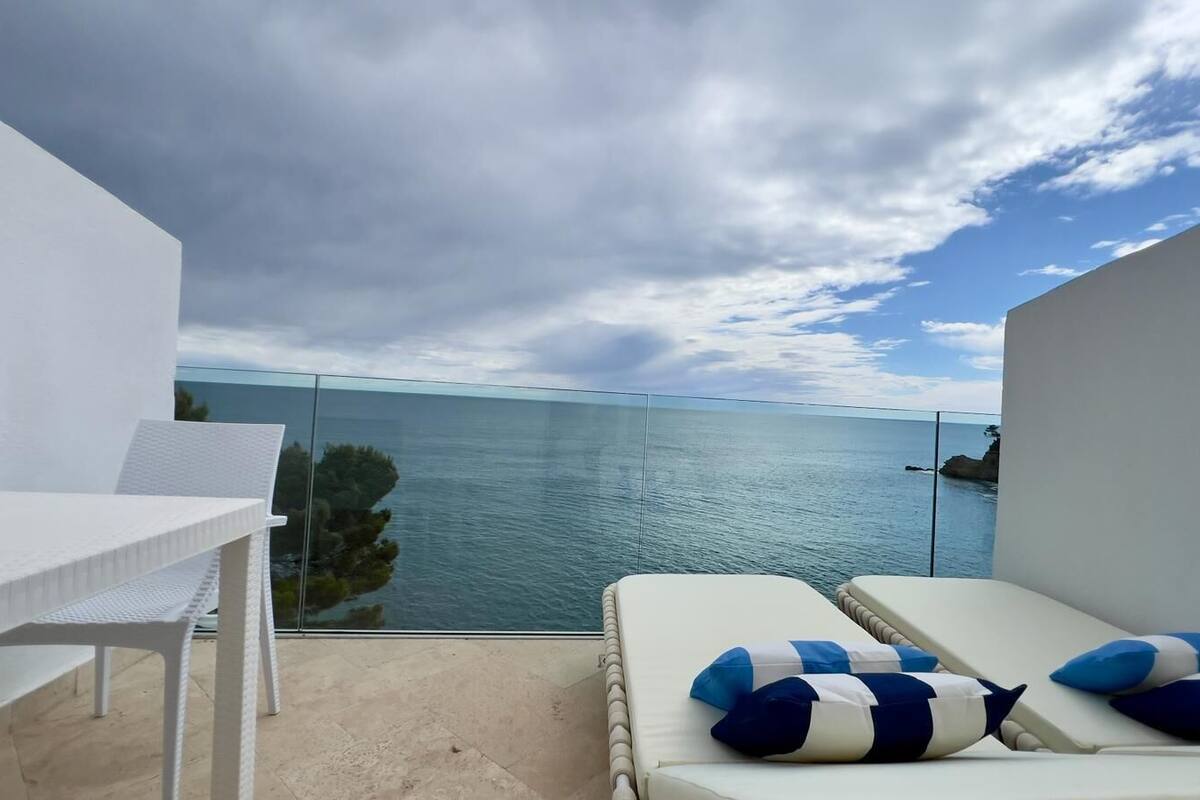 Un balcón con una mesa y sillas blancas, una zona de estar con cojines y vista al mar.