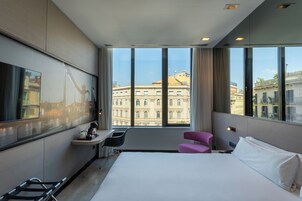 Une chambre d’hôtel avec un grand lit, un bureau, une chaise et une vue sur les bâtiments de la ville, visible à travers de grandes fenêtres.