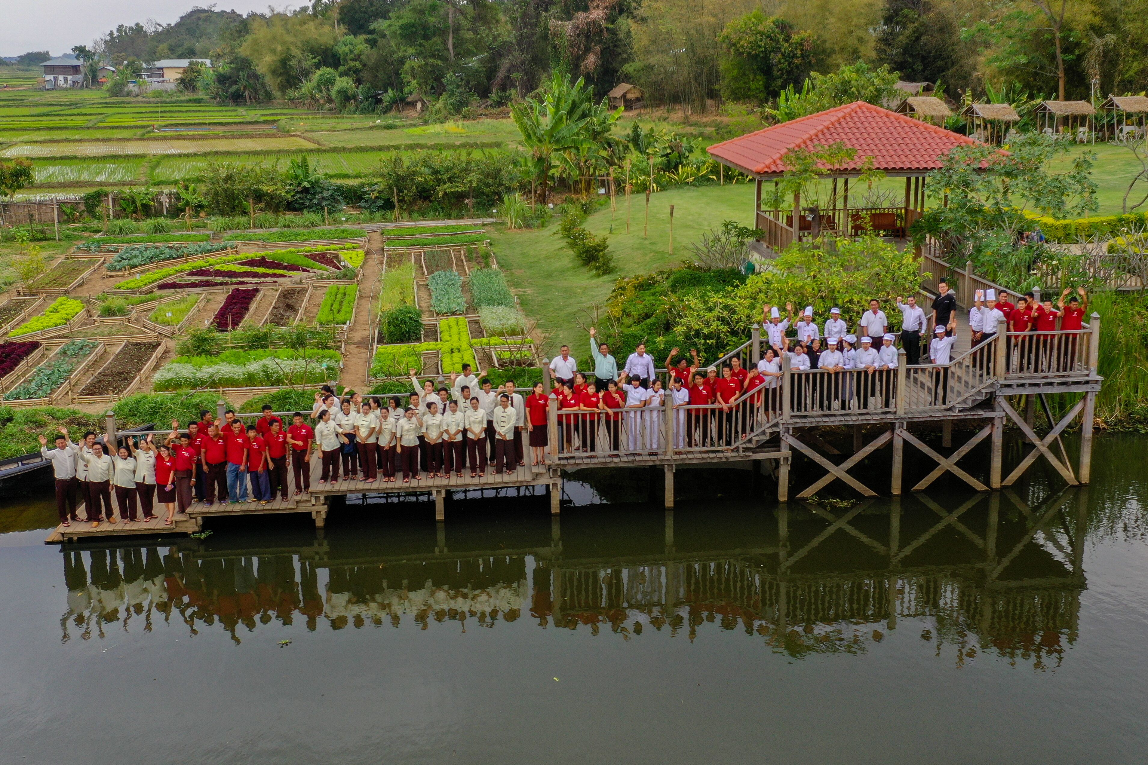 Un grupo de personas en un puente de madera sobre un cuerpo de agua, con un jardín y una estructura de techo rojo al fondo.