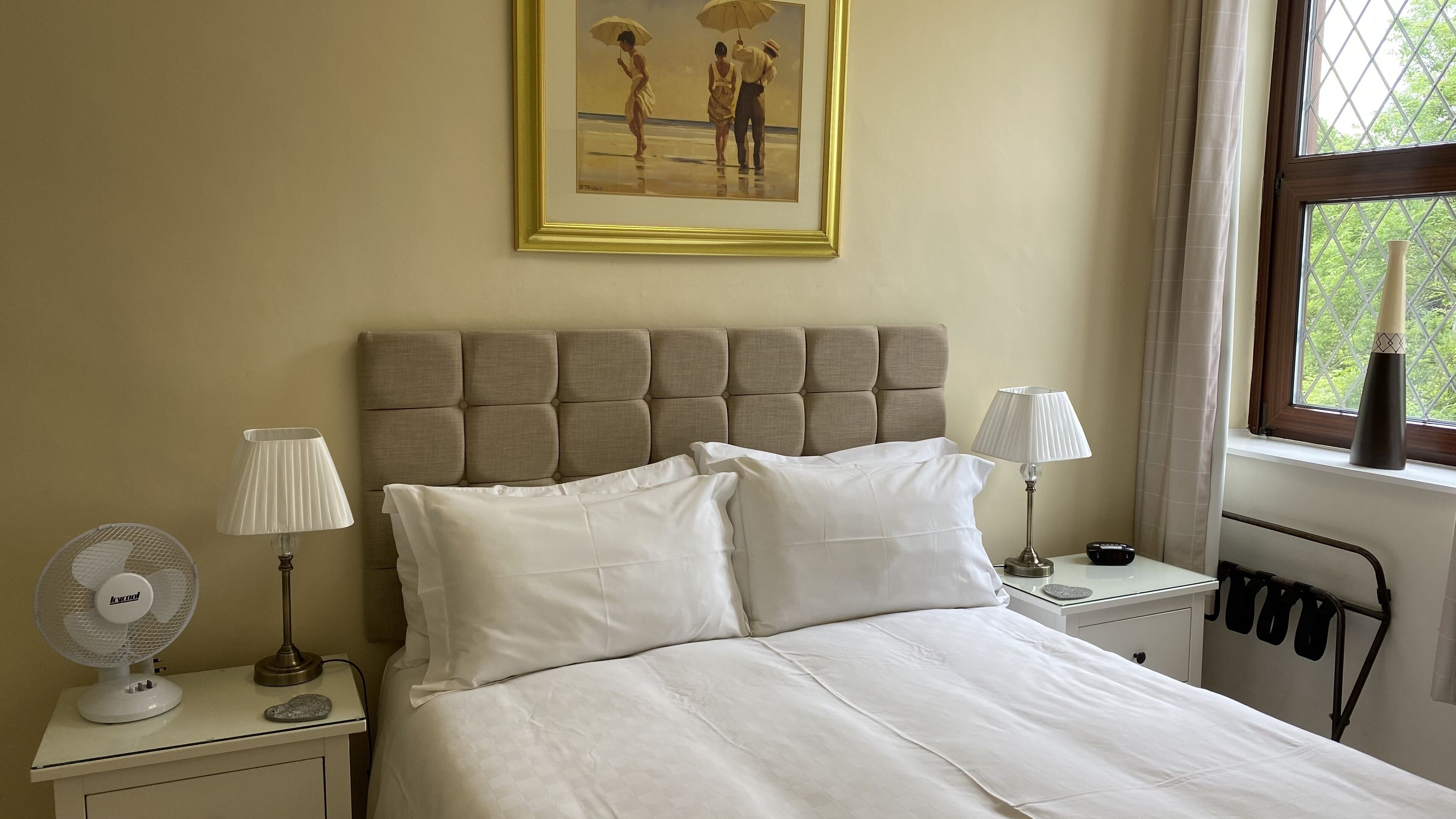 A neatly made bed with white linens, a headboard, two bedside lamps, a fan, and a painting above the bed.