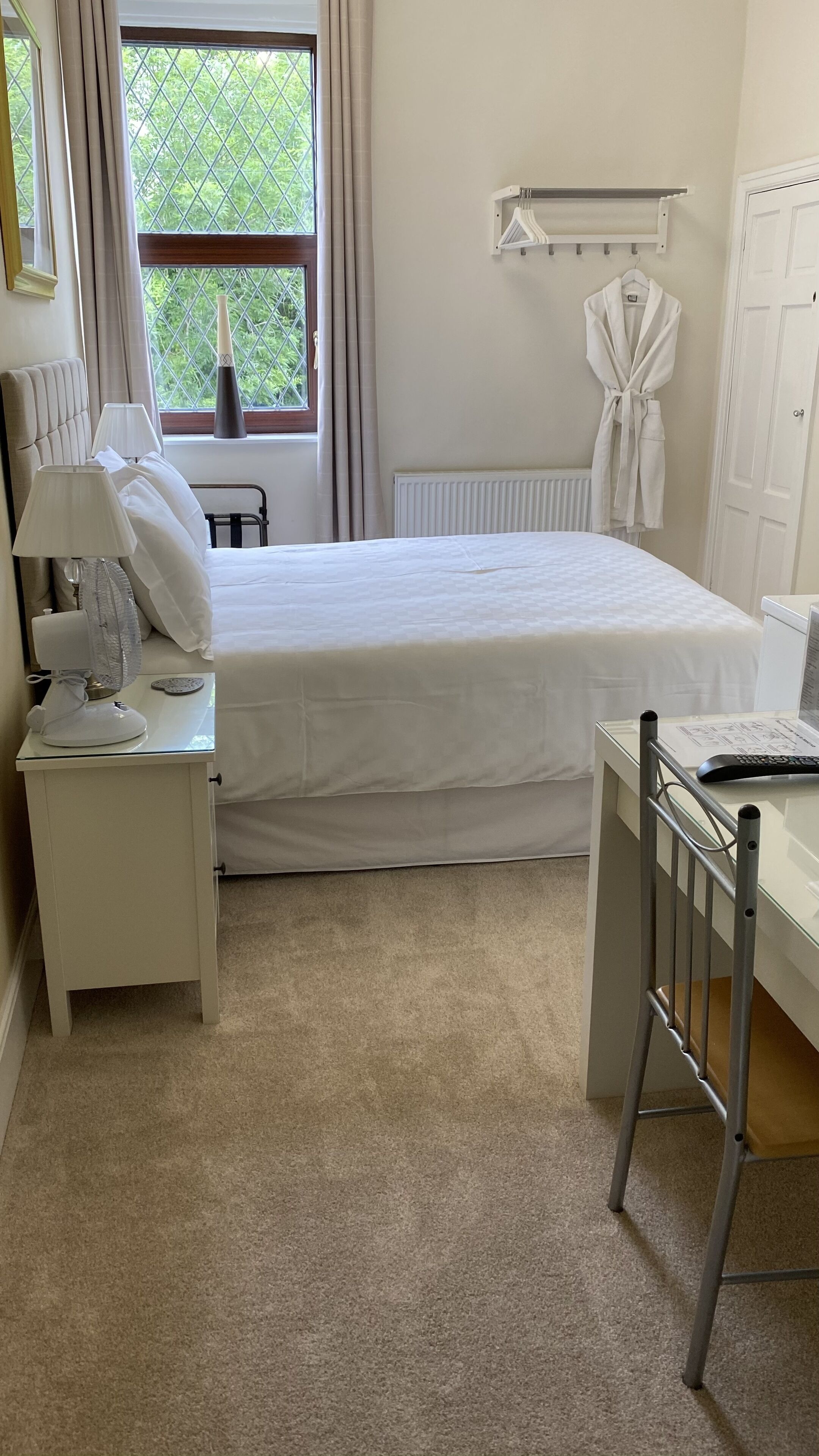 A hotel room with a bed, bedside table with a lamp and fan, a radiator, a robe hanging on the wall, and a desk with a chair.