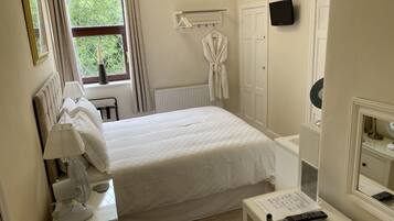 Un dormitorio ordenado con cama, lámparas de noche, un radiador, una bata colgada en un perchero, un televisor y un espejo.