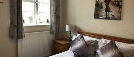 Una camera da letto con un letto, un comodino, una lampada, una finestra con tende e un quadro appeso al muro.