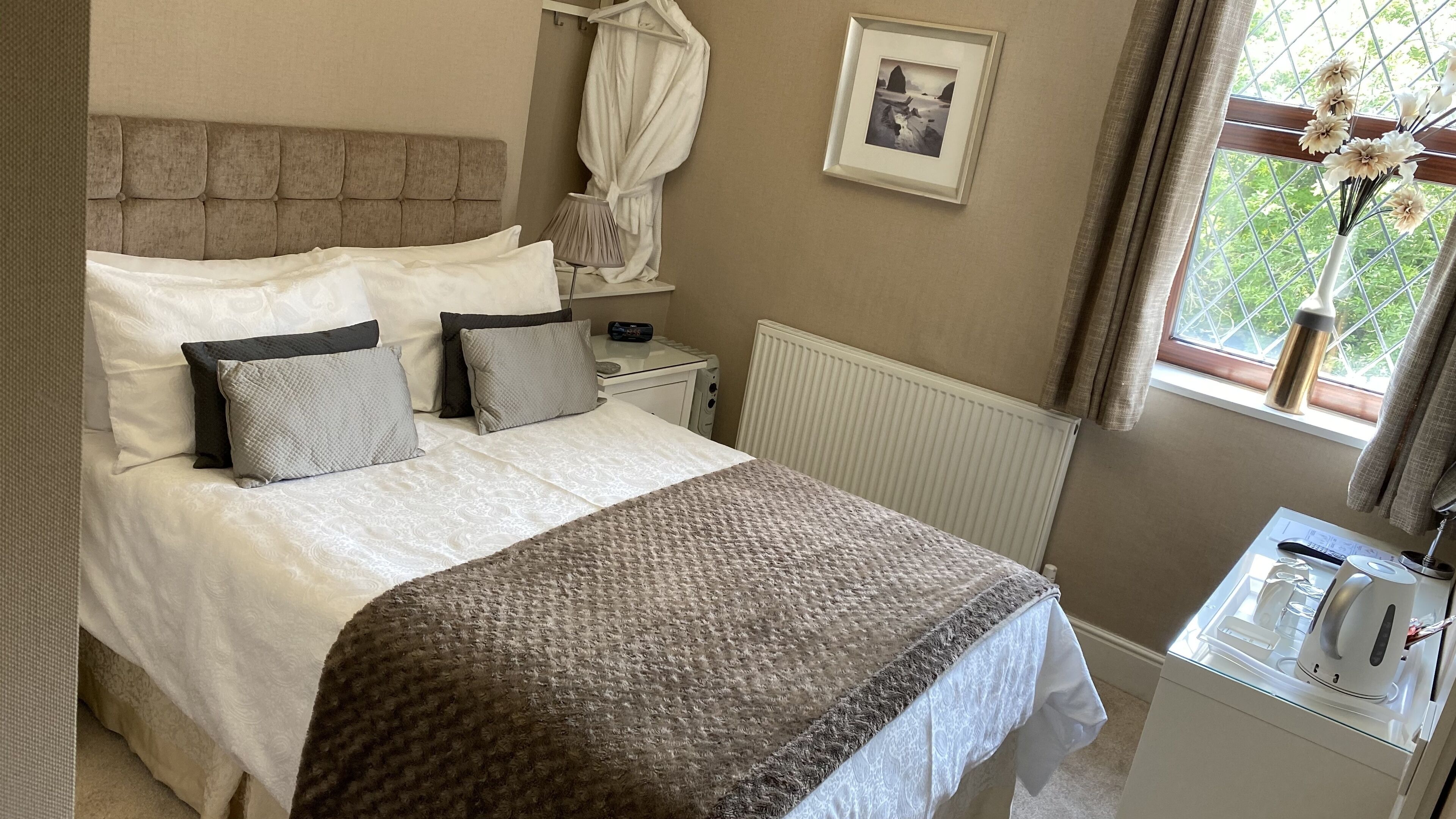 A neatly made bed with a patterned blanket, a bedside table with a lamp, a framed picture on the wall, and a window with curtains.