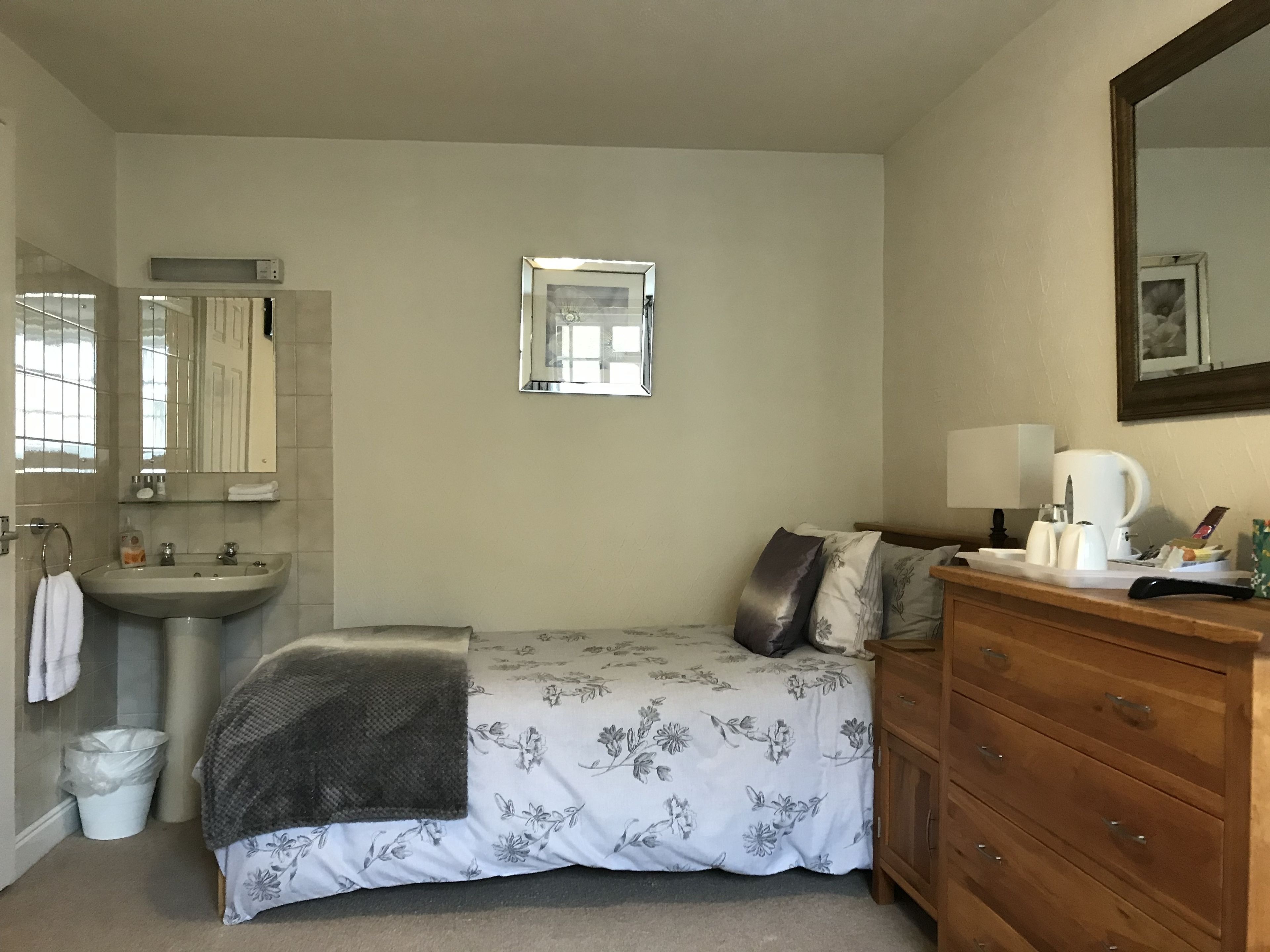 Un dormitorio con cama, mesita de noche con lámpara, cómoda, espejo y baño con lavamanos y perchero para toallas.