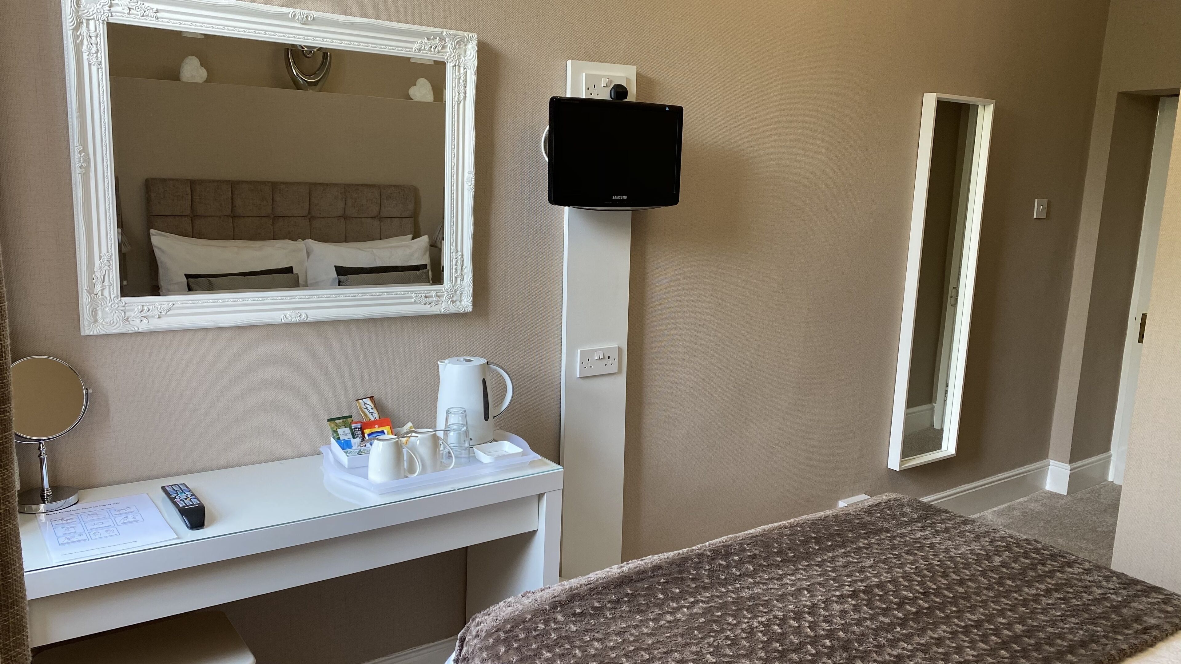 Habitación de hotel con cama, mesita de noche con control remoto, espejo, cafetera y televisor de pantalla plana montado en la pared.