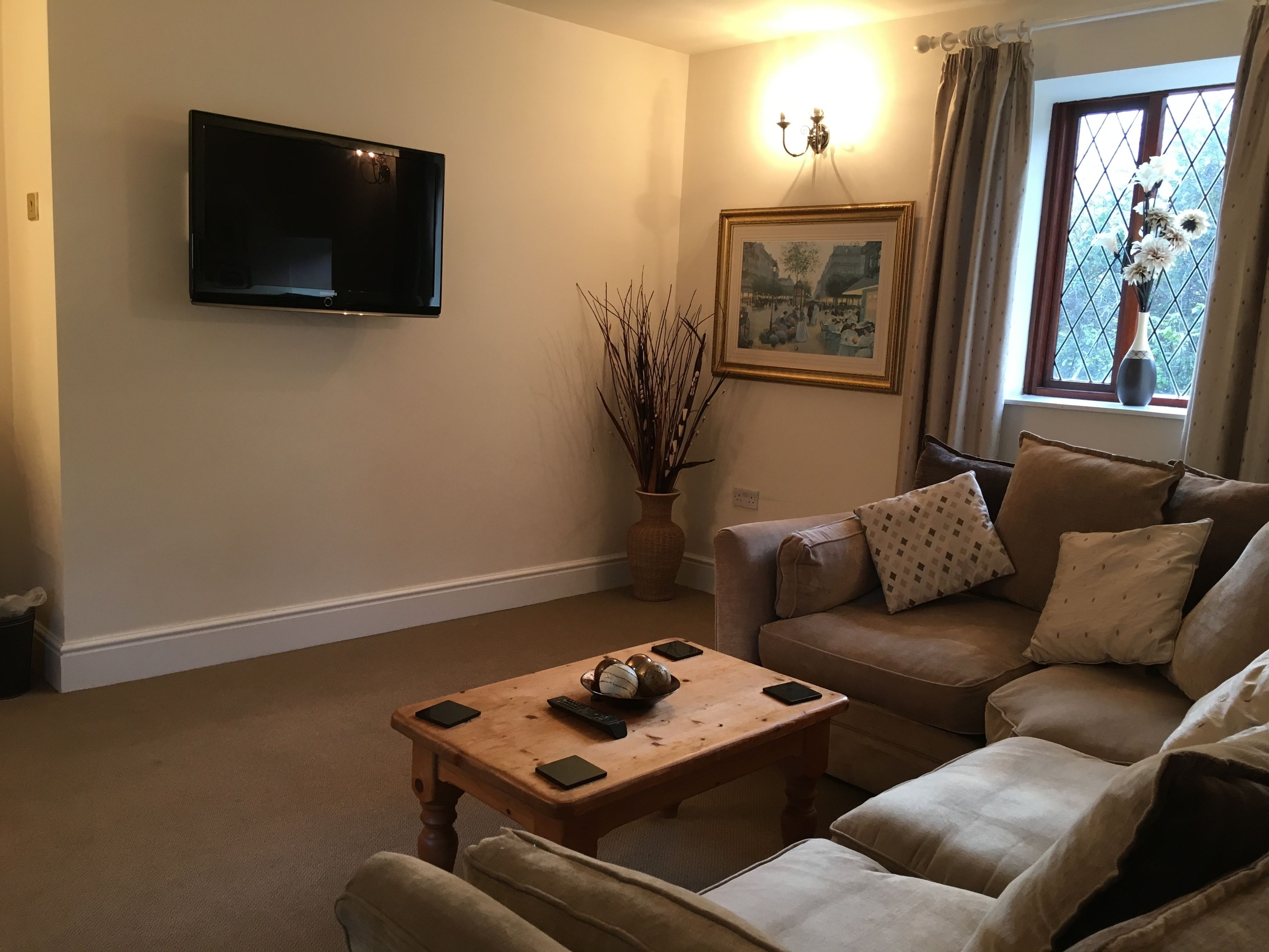 A living room with a flat-screen TV mounted on the wall, a wooden coffee table with remote controls, a beige sofa with patterned cushions, and a framed picture above the sofa.