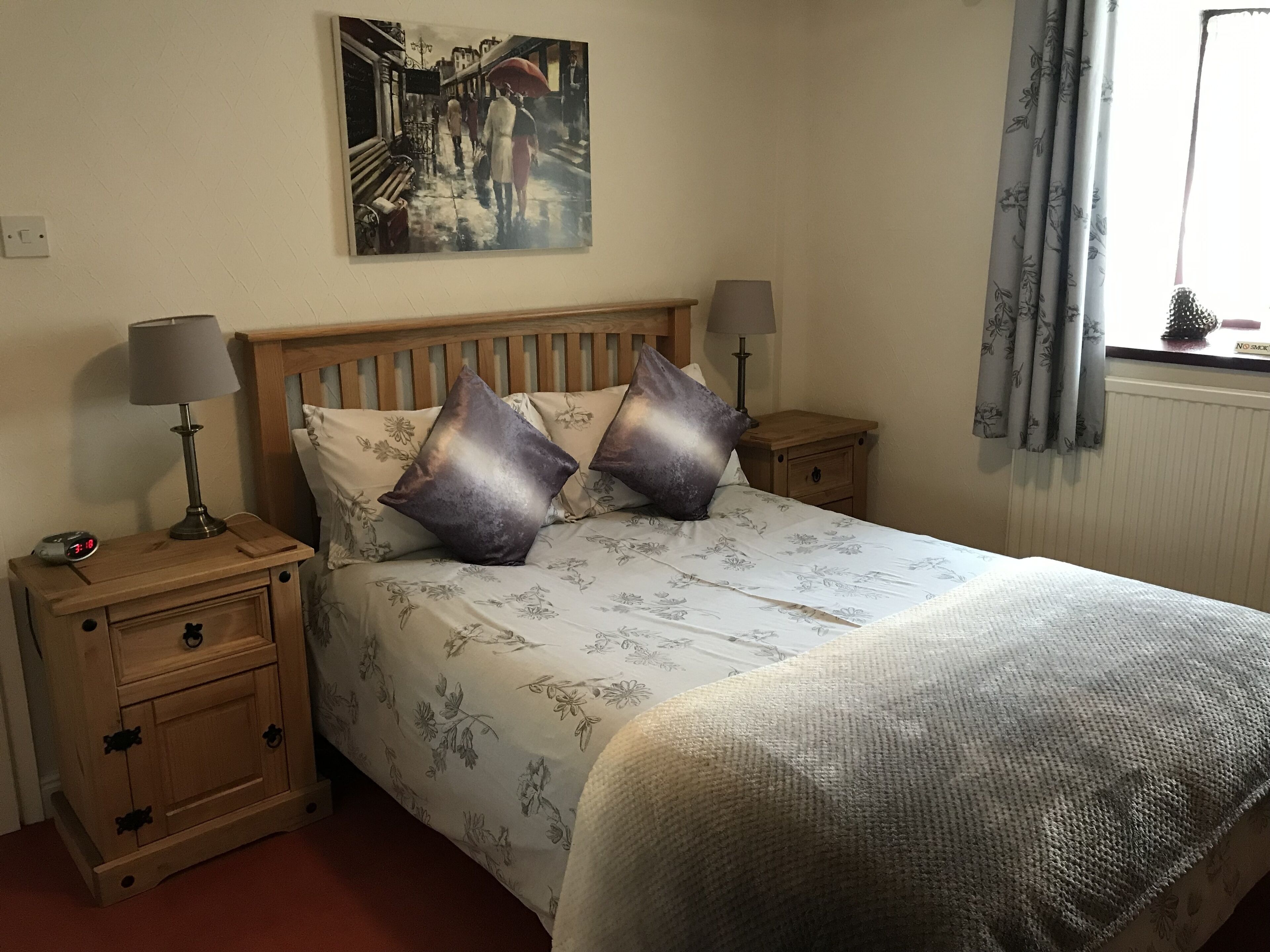 A bedroom with a wooden headboard, floral bedding, two bedside tables with lamps, and a painting on the wall.