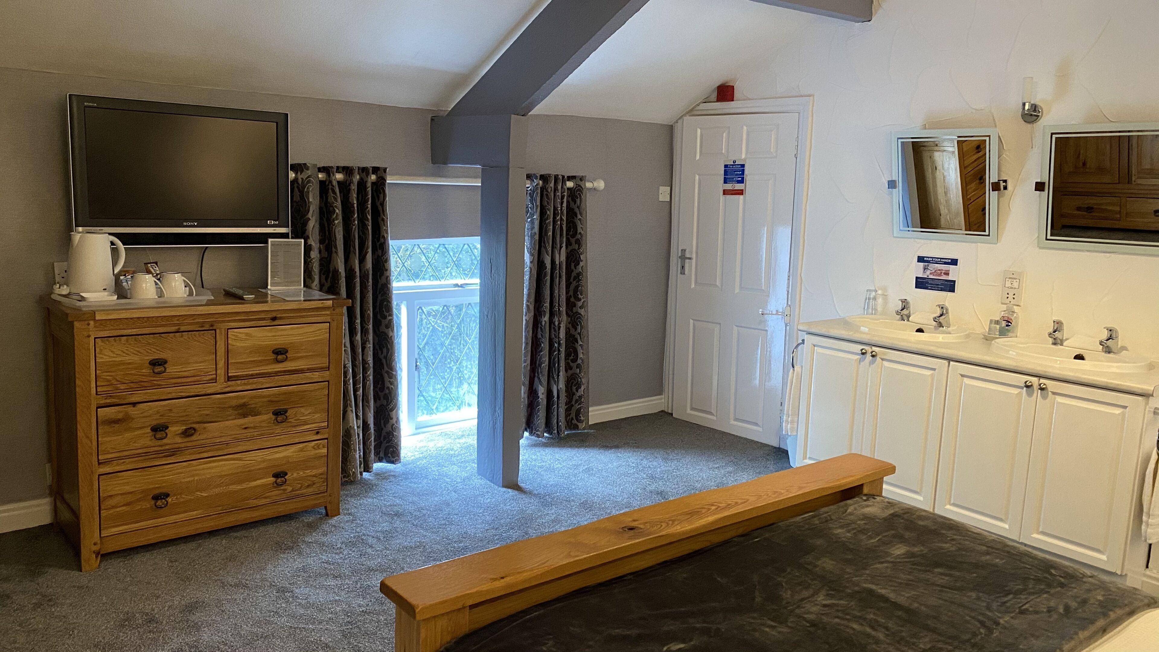 A hotel room with a wooden dresser, a bench, a television, a bathroom with a sink and mirror, and a door leading to a balcony.