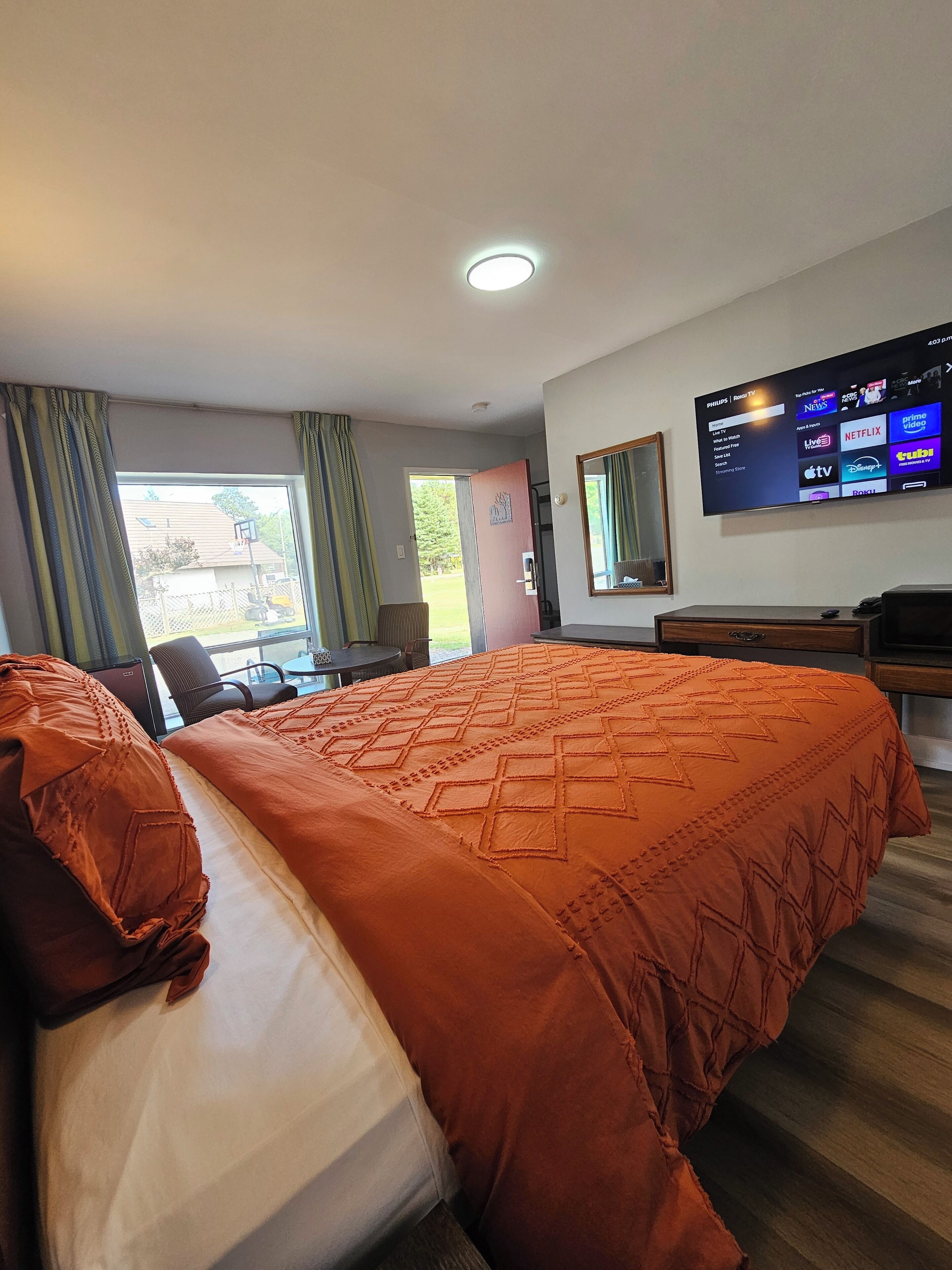 Ein Hotelzimmer mit einem großen Bett, einem Schreibtisch, einem Fernseher, der Senderauswahl zeigt, und einem Fenster mit Vorhängen.