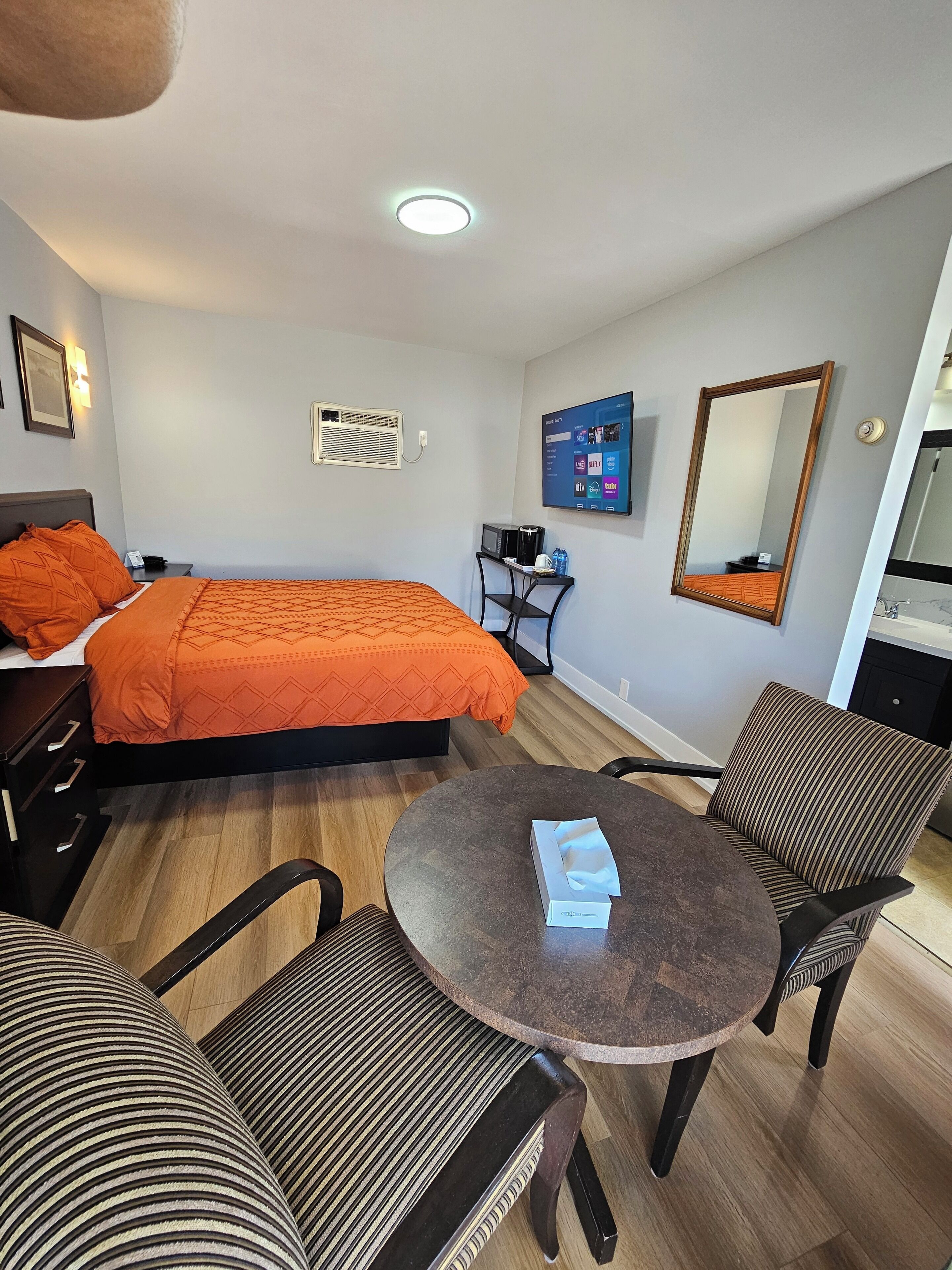 A hotel room with a bed, a desk, a chair, a round table, and a large mirror.